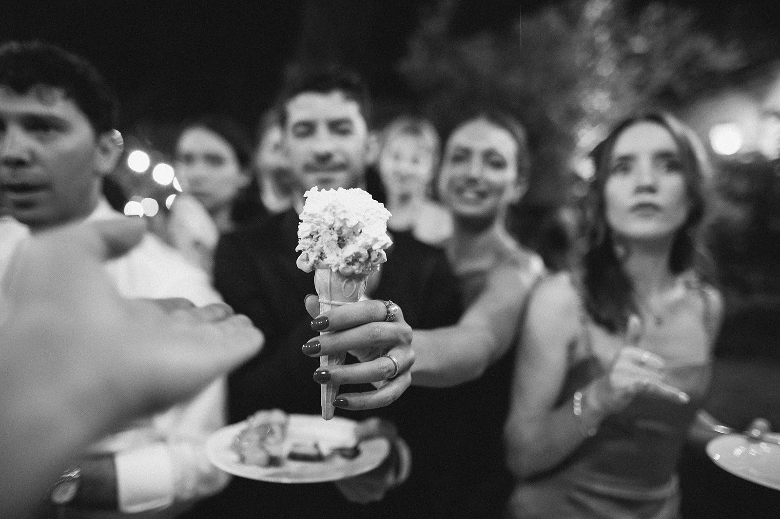 Black and white photo of a group of people at a social gathering, with a woman in the center holding a small ice cream cone with what appears to be whipped cream on top.