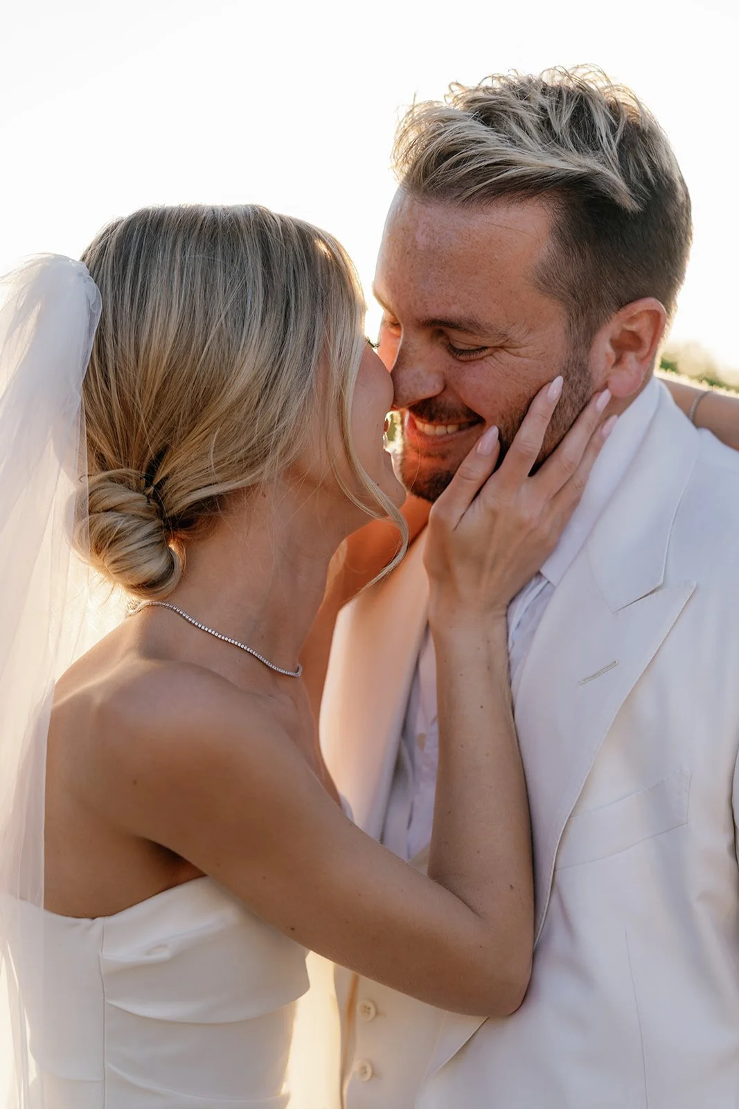 A bride and groom share a joyful kiss during their wedding, with the bride gently holding the groom's face and the groom smiling with eyes closed in a close-up shot at sunset.