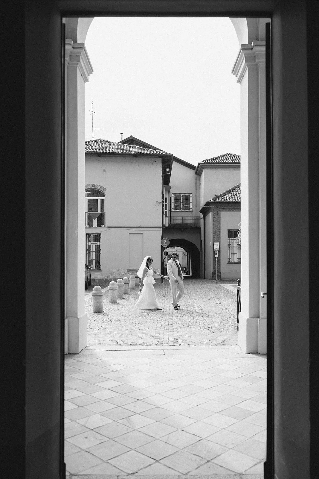 A couple dressed in wedding attire walking hand in hand through an arched doorway into a cobblestone street with historic buildings.