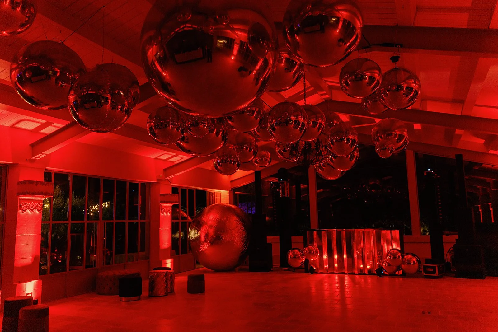 A room decorated with numerous large shiny red metallic balloons hanging from the ceiling, with some smaller balloons on the floor. The room is lit with red lighting, creating a vibrant and festive atmosphere.