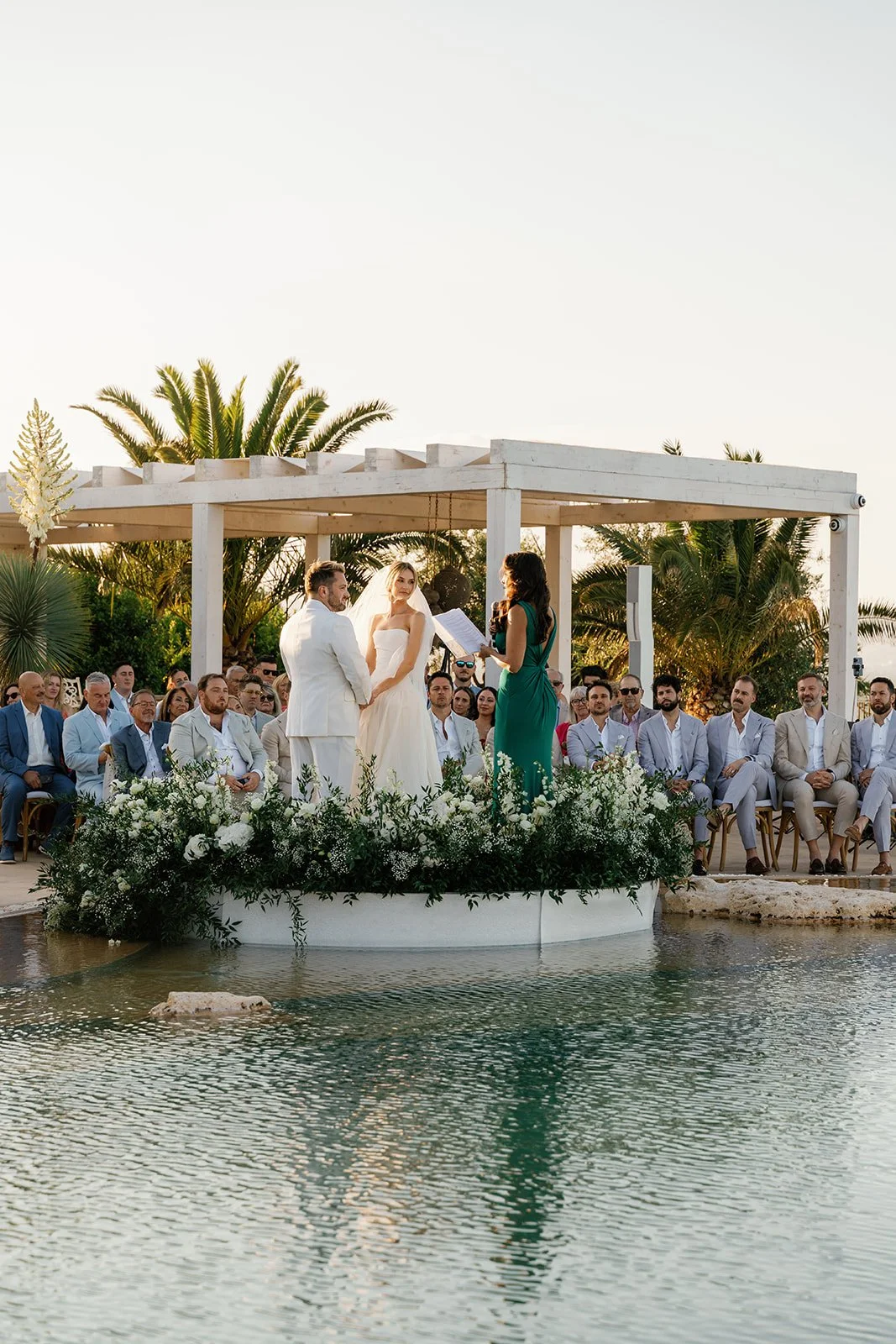 A couple getting married during an outdoor ceremony by a pool, with guests seated around them, officiant reading from a paper, and palm trees in the background.