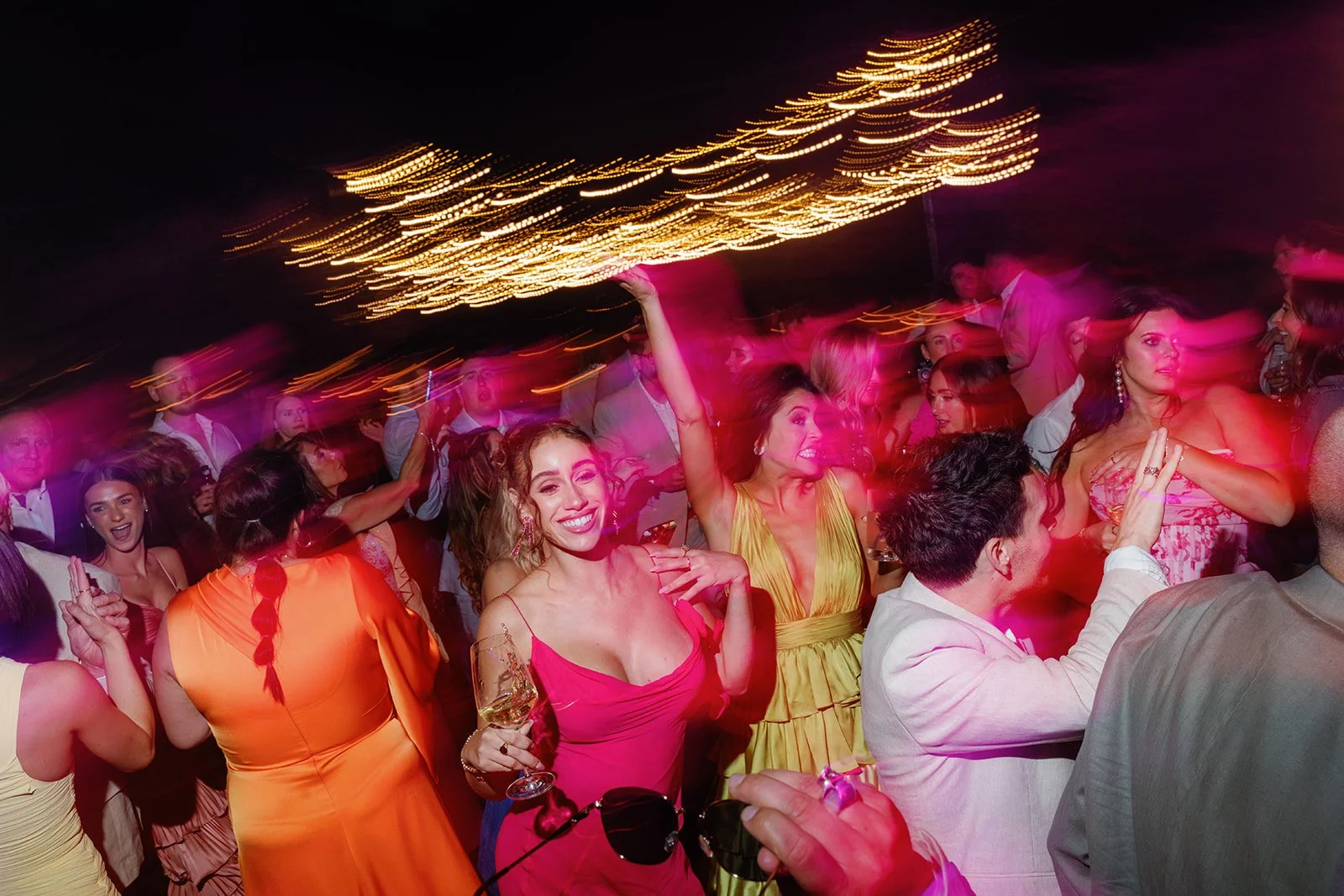 A lively party scene with women dancing, smiling, and holding drinks, illuminated by colorful lighting and string lights overhead.