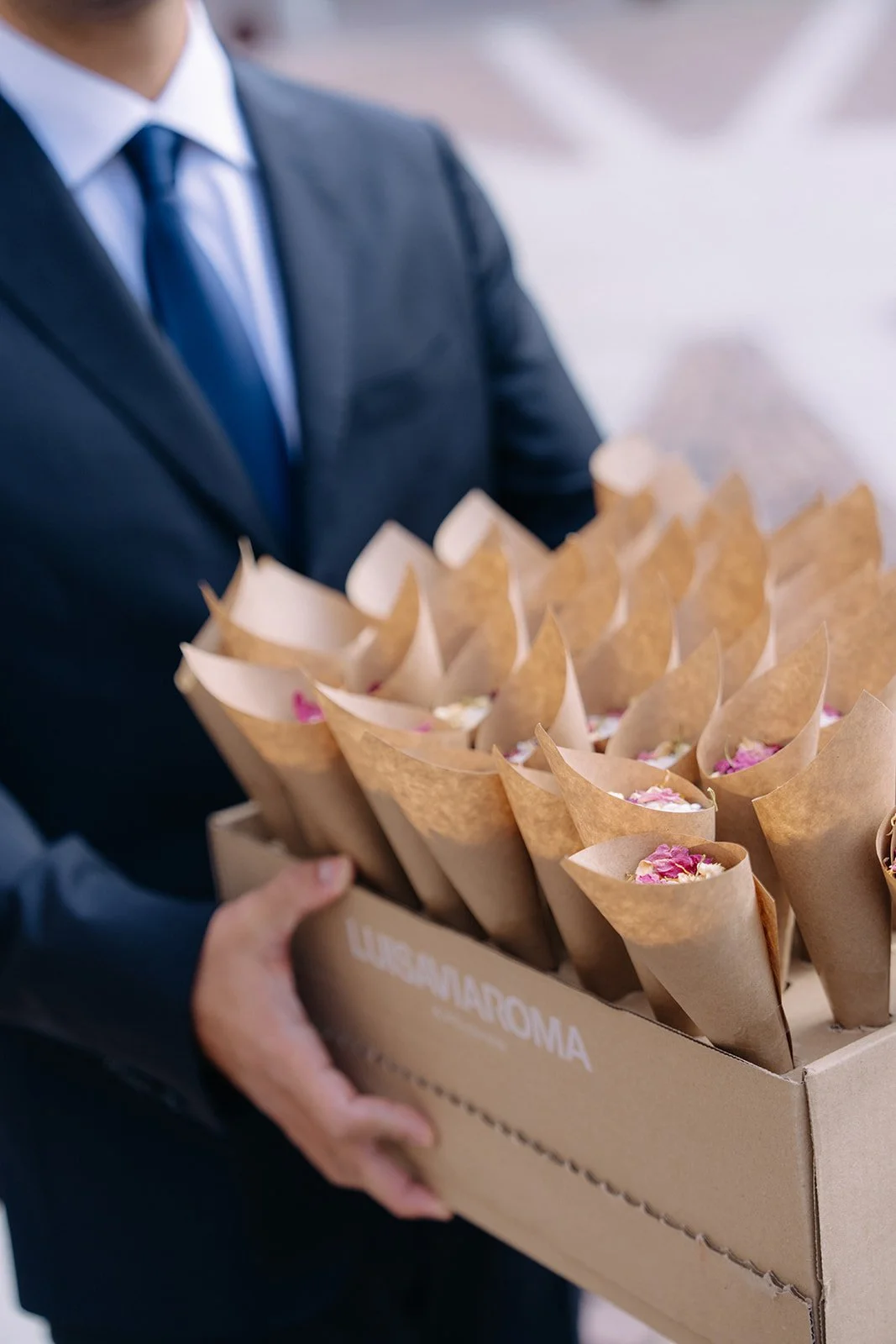 A man in a suit holding a cardboard box filled with small cone-shaped packages wrapped in brown paper, topped with pink and white flower petals.