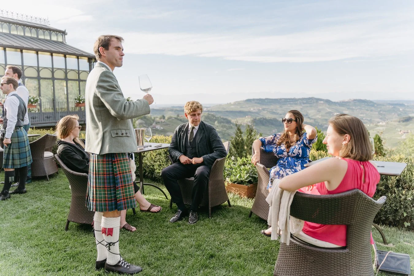 People socializing outdoors on a patio with scenic hills in the background. One man in traditional Scottish attire, including a kilt, holding a wine glass, stands near seated women and men dressed in casual and semi-formal clothing.