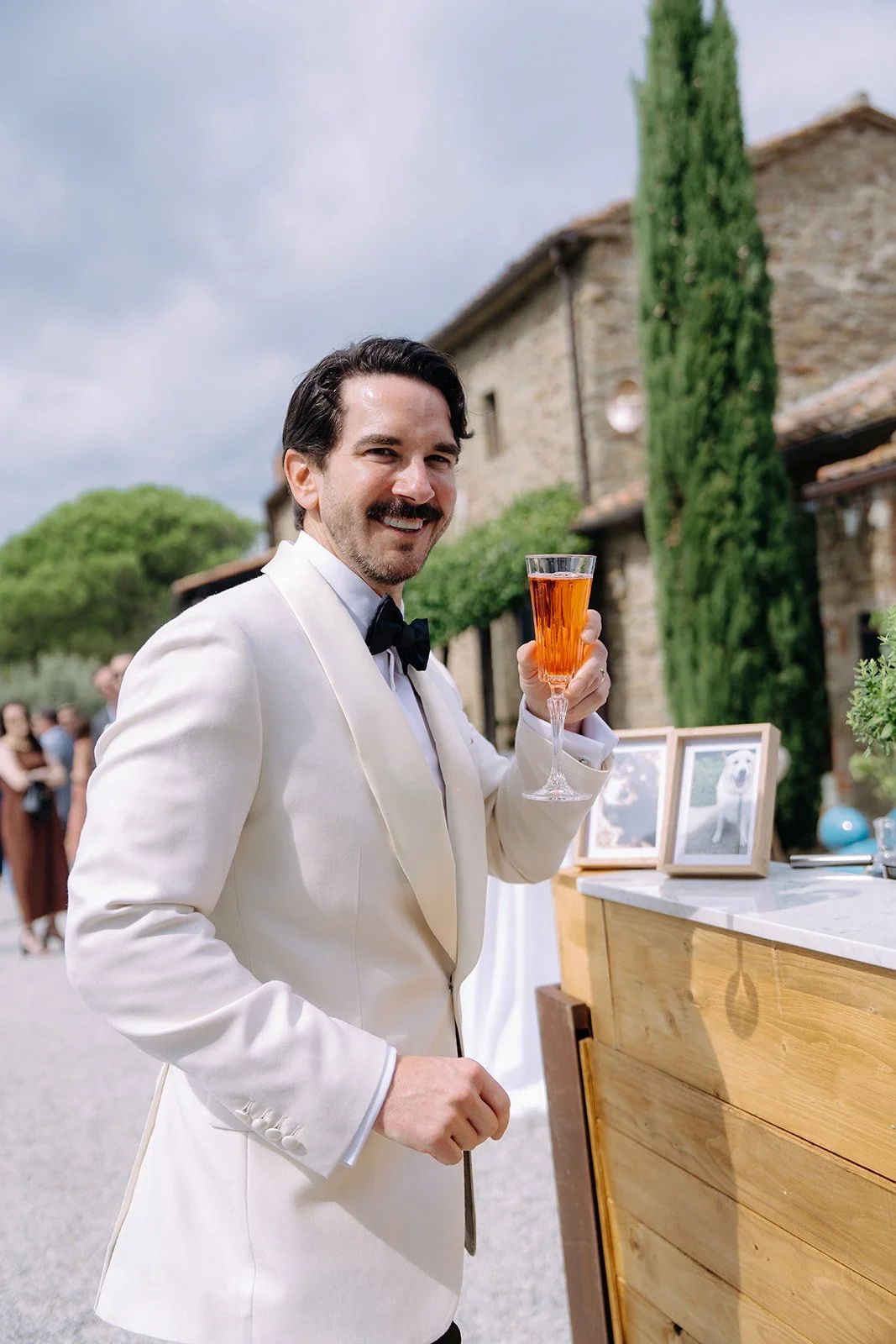 A man in a white tuxedo with a black bow tie, smiling and holding a glass of pink-colored beverage, standing outdoors with a rustic stone building and green trees in the background.