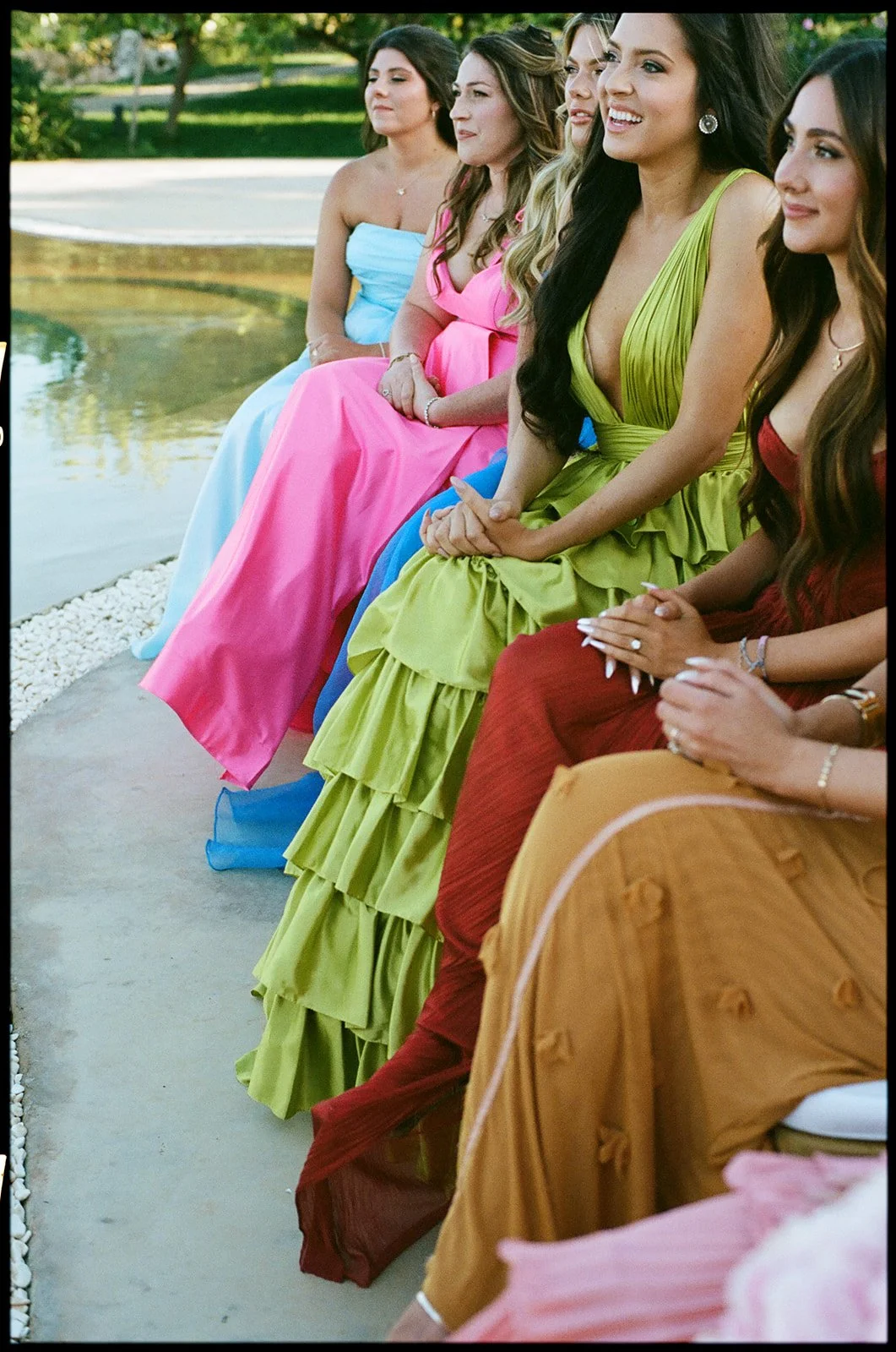 A group of women sitting outdoors by a pond, dressed in colorful gowns, smiling and enjoying a social event.
