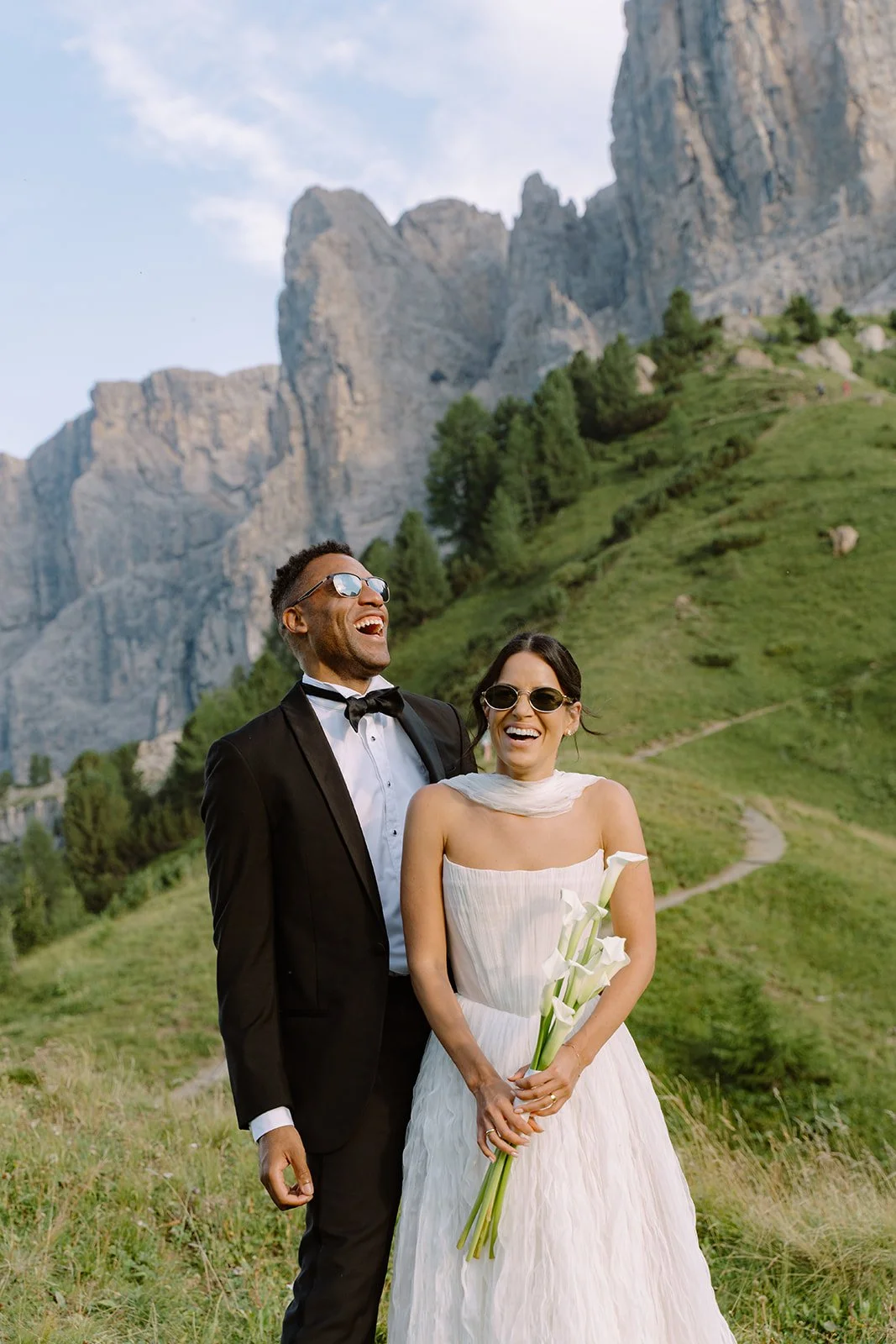 A happy couple in wedding attire standing outdoors with mountains and green hills in the background. The man is wearing a black tuxedo with a bow tie and sunglasses, and the woman is in a strapless white wedding dress with sunglasses and holding a bo