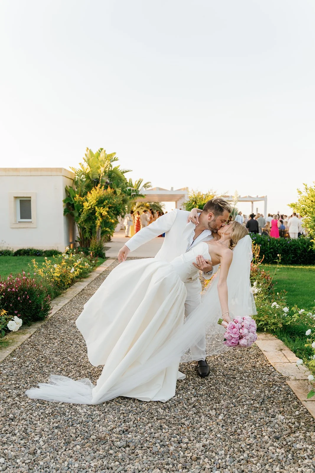 A newlywed couple shares a romantic kiss outdoors at their wedding reception. The groom dips the bride while she holds a bouquet of pink flowers, with guests in the background enjoying the celebration.