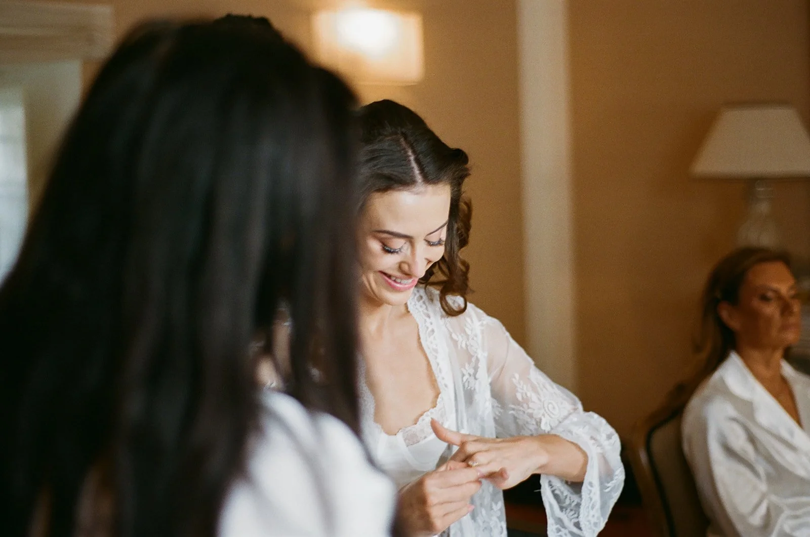 Three women sitting in a room, with one woman smiling and looking at her hand, dressed in a white lace robe, while another woman with long black hair and a third woman with blonde hair sit nearby.