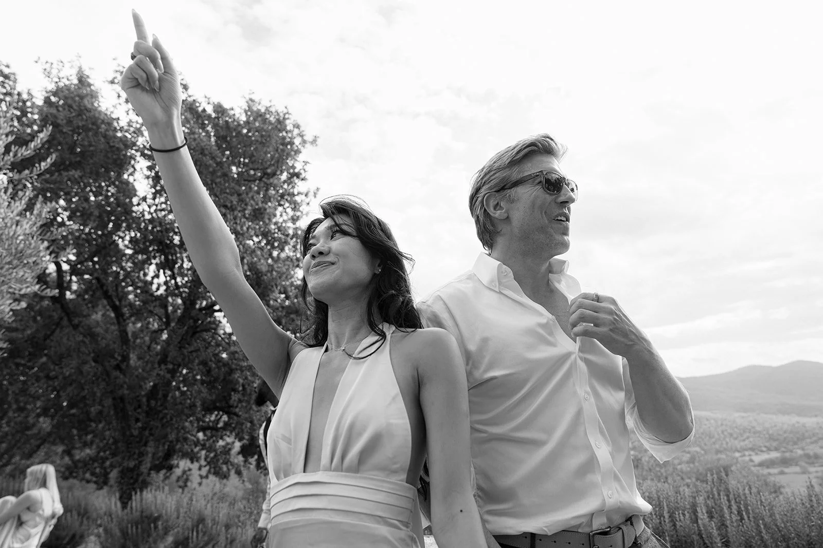 A woman and a man standing outdoors, with the woman raising her arm and pointing upward while the man looks to the right, both smiling. The image is in black and white, with trees and open landscape in the background.
