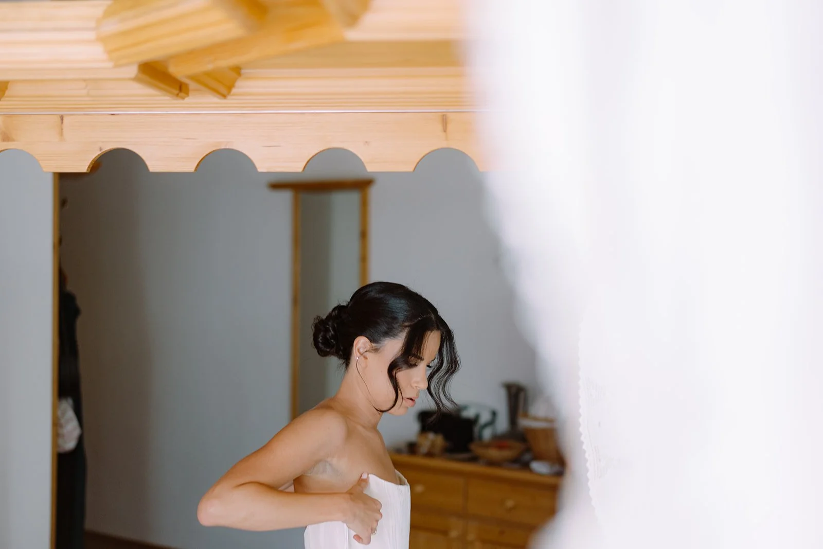 A woman wrapped in a white towel, adjusting it on her shoulder, standing in a room with wooden ceiling and furniture, with a partial view of a mirror and some objects in the background.