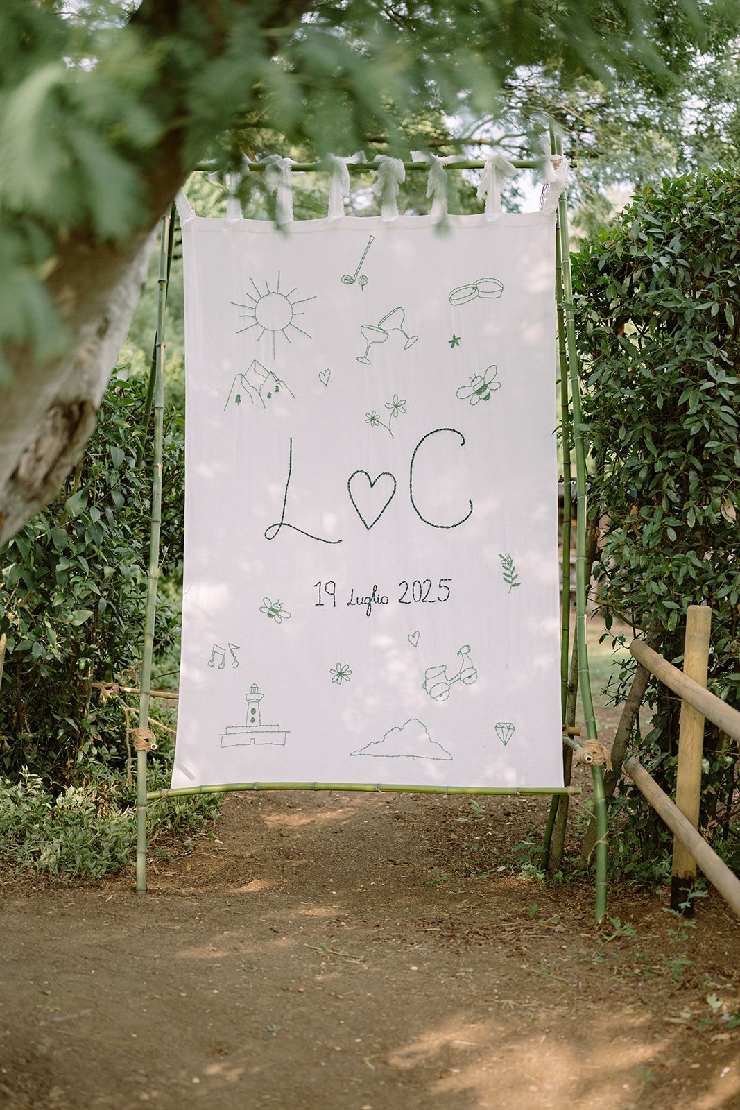 A white fabric banner hanging outdoors among trees and bushes with doodles of the sun, mountains, a lighthouse, flowers, bees, bikes, rings, musical notes, clouds, and various symbols. The banner has the text "L♥C" and "19 Luglio 2025" written on it.