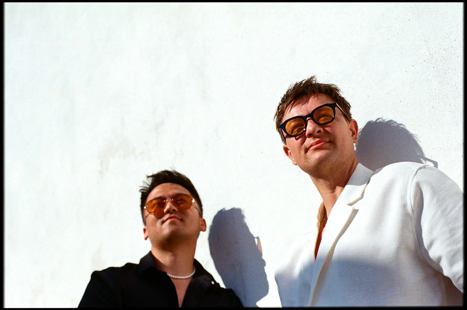 Two men with sunglasses leaning against a white wall, casting shadows. One is wearing a black shirt and pearl necklace, the other a white jacket.