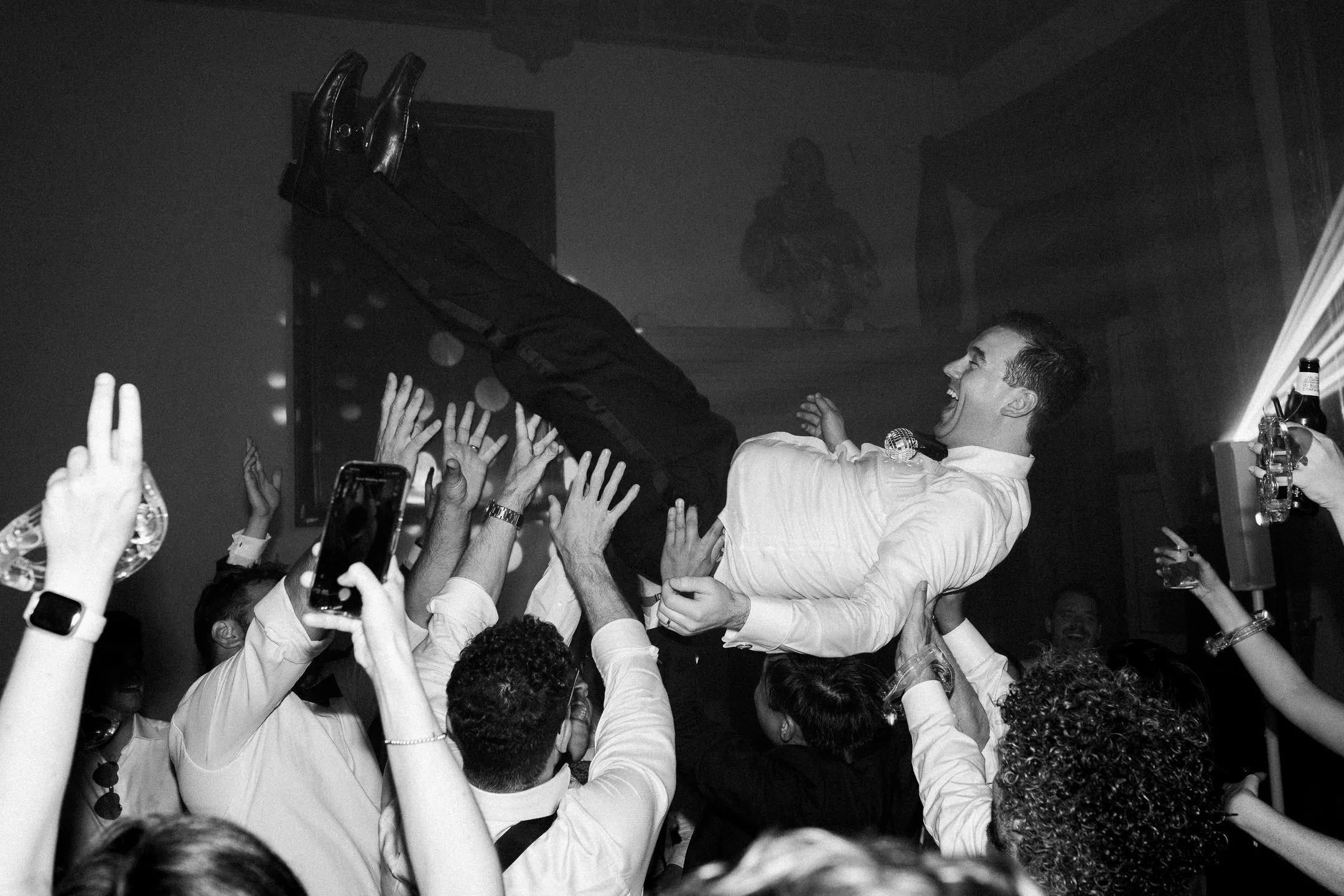 A young man in a white shirt is lifted above a crowd at a party, smiling and laughing, with people reaching out to support him. Some are taking photos and making peace signs.