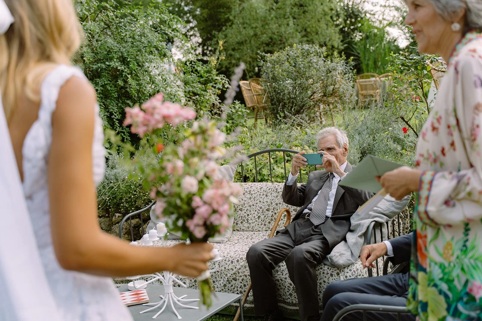 An elderly man in a suit is sitting on a garden bench, taking a photo with his smartphone of a woman holding a large bouquet of flowers. Two other women are in the scene, one on the left holding a bouquet, and another on the right holding papers, all