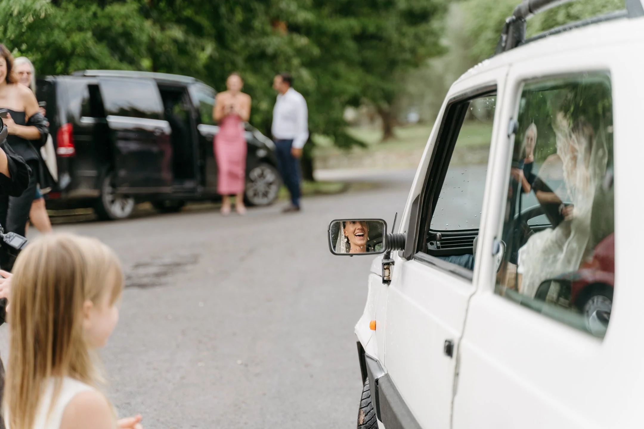 A woman in the side mirror of a white vehicle is smiling and laughing. People are gathered around cars in a park with trees in the background.