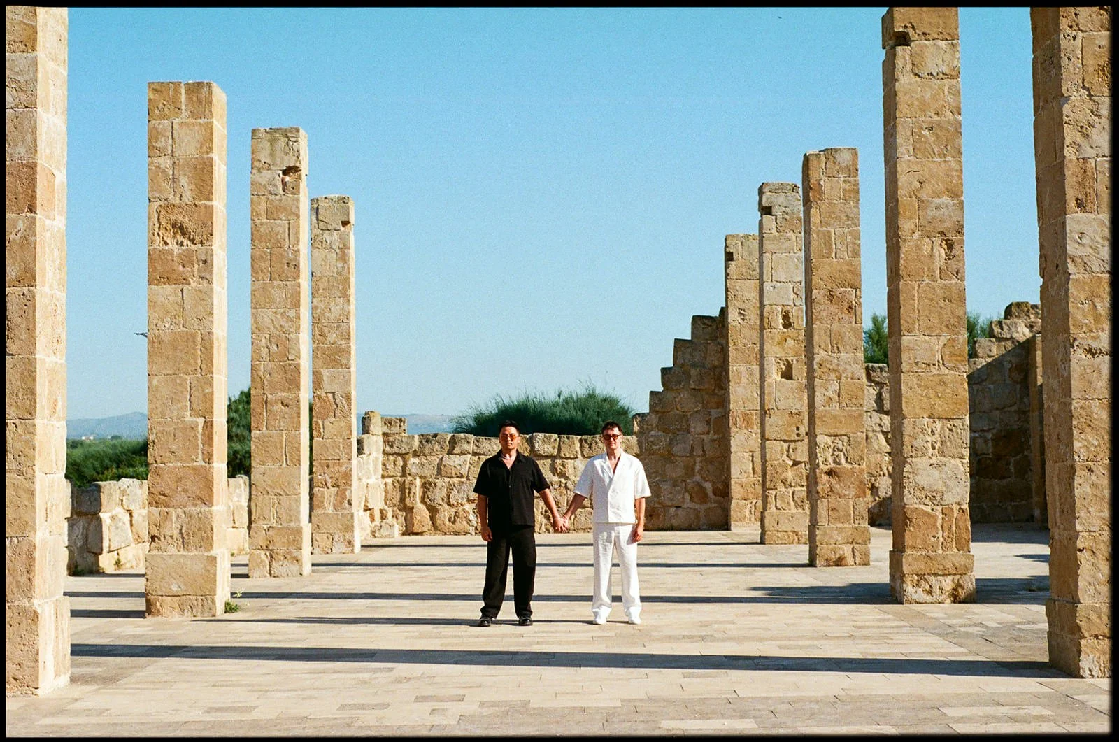 Two people holding hands and standing in the center of ancient stone ruins against a clear blue sky.