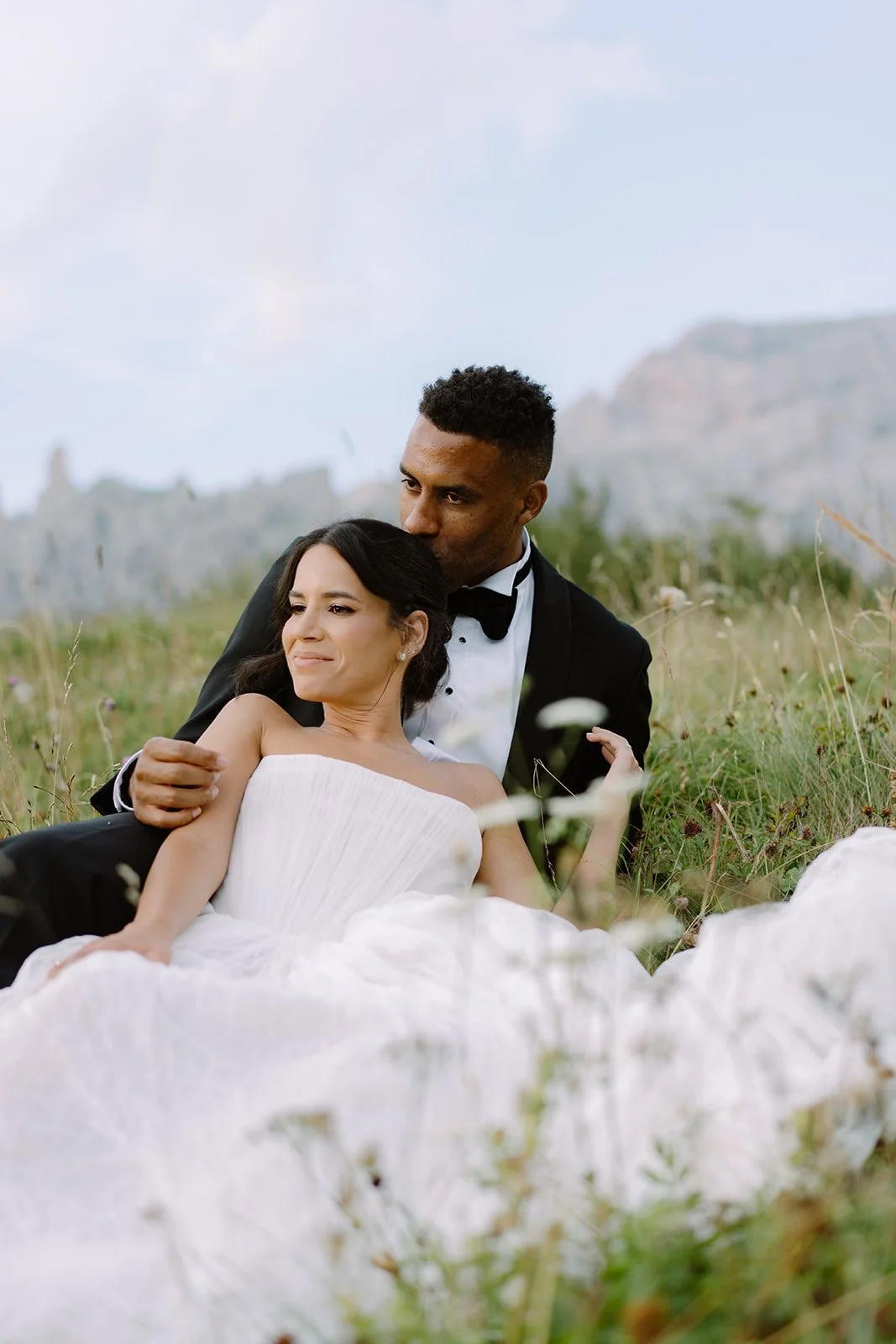 A couple in wedding attire sitting in a grassy field, with the man kissing the woman's forehead.
