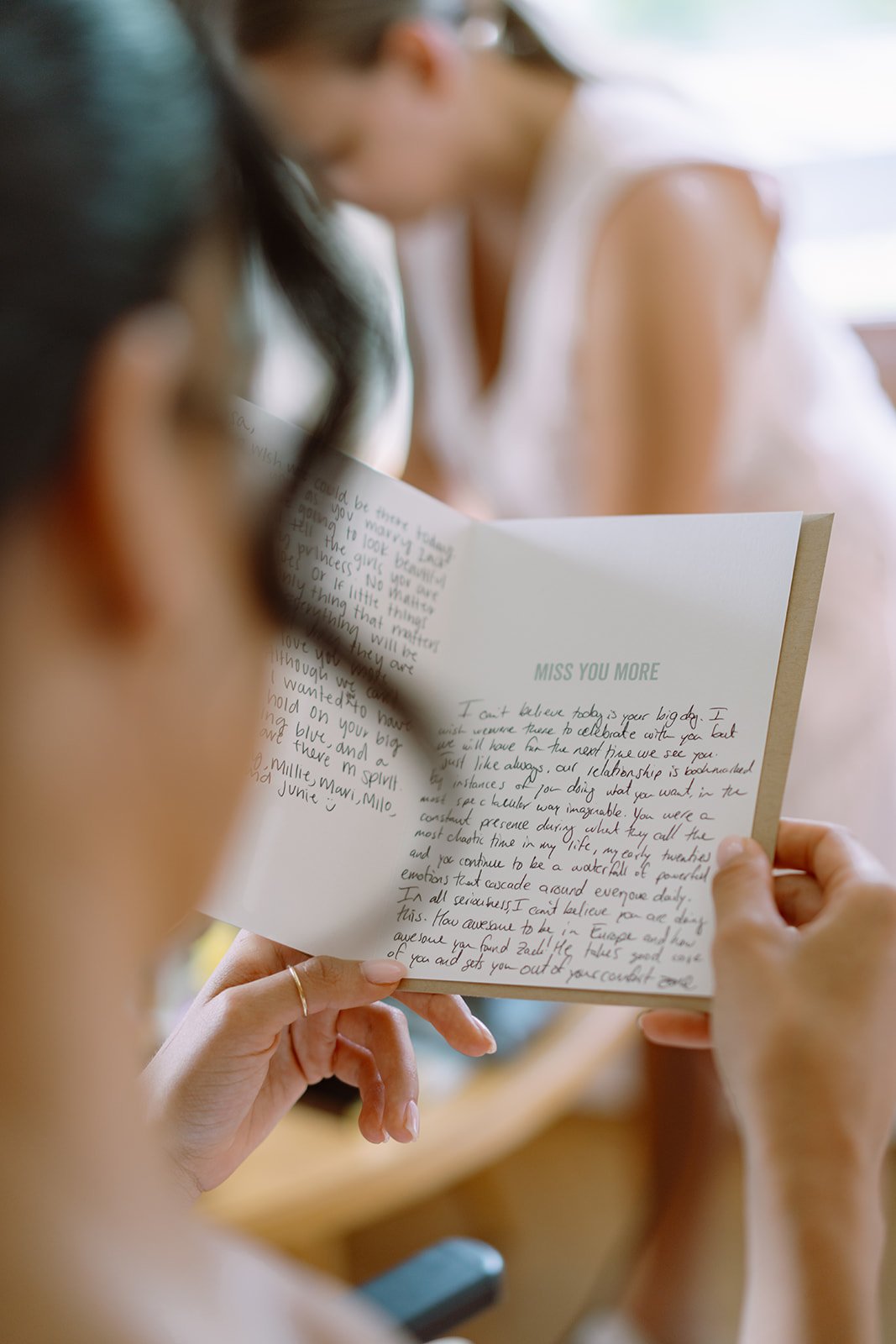 A woman is reading a handwritten letter in a greeting card. The letter is titled 'MISS YOU MORE' and the woman has long hair, wearing a ring, and is focused on the message.