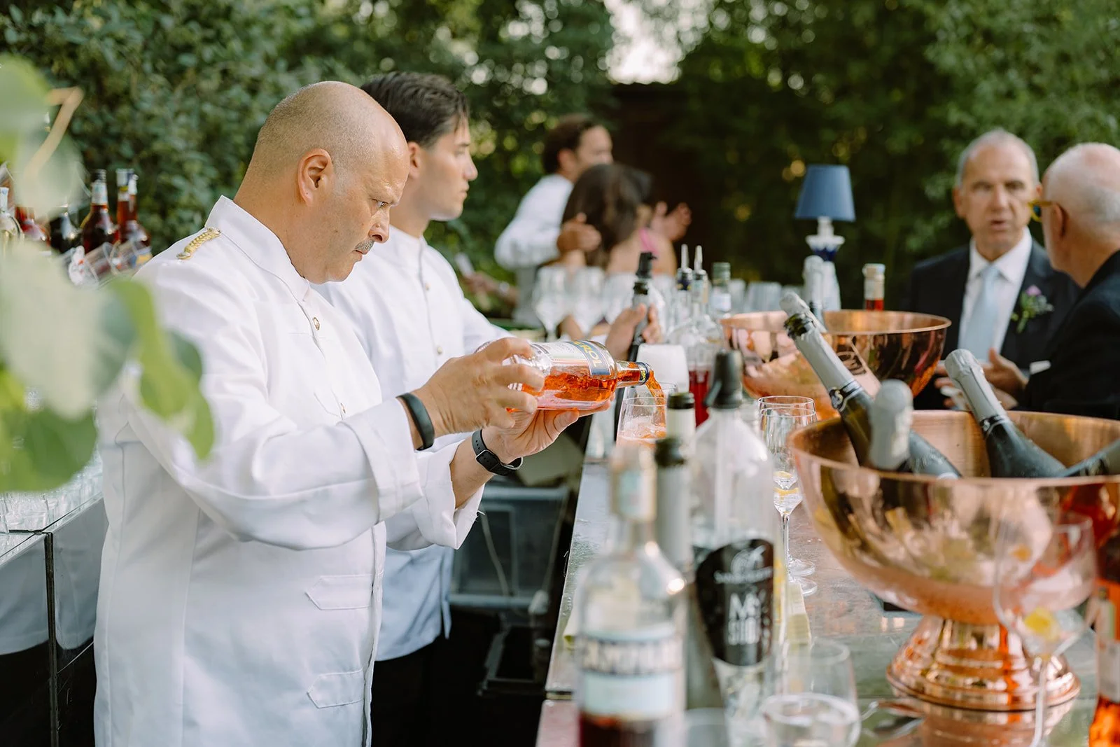 Chef pouring pink beverage at outdoor bar during event with guests.