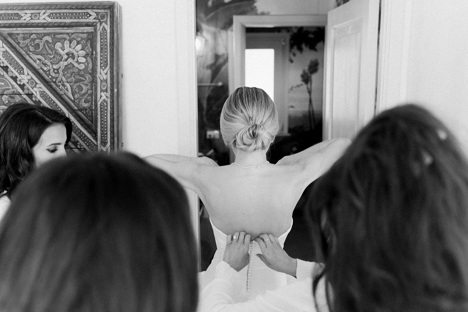 A bride with her back to the camera, wearing a strapless wedding gown, is being helped into her dress by two women while another woman looks on, in a room with art on the wall and a window showing palm trees outside.