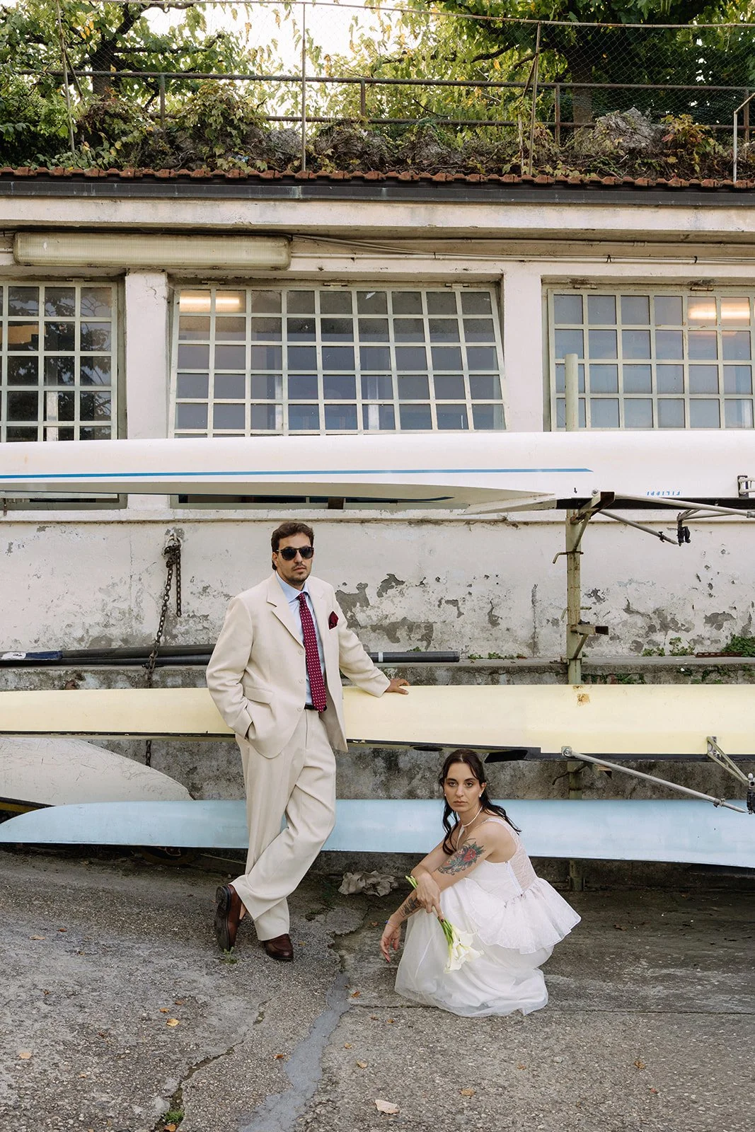 A man in a beige suit and sunglasses standing next to a woman in a white dress holding a white flower in front of a wall with stacked boat hulls and windows.