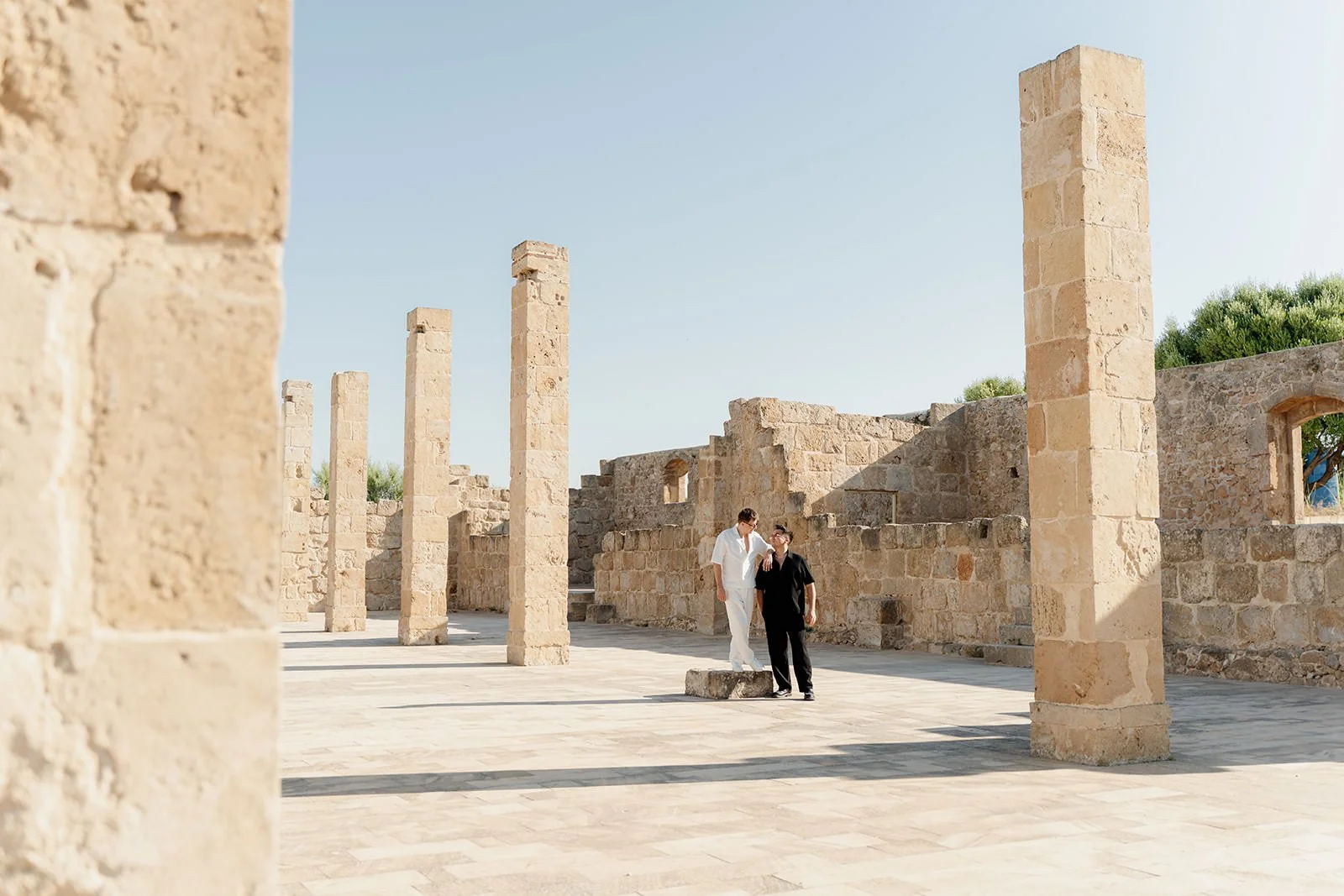 Two people walking together through ancient stone ruins with tall stone columns, clear sky, and some greenery in the background.