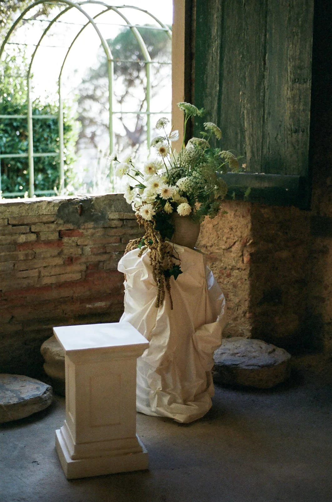 A bouquet of white flowers in a vase, draped with a white cloth, placed on a pedestal inside a rustic room with a window showing greenery outside.