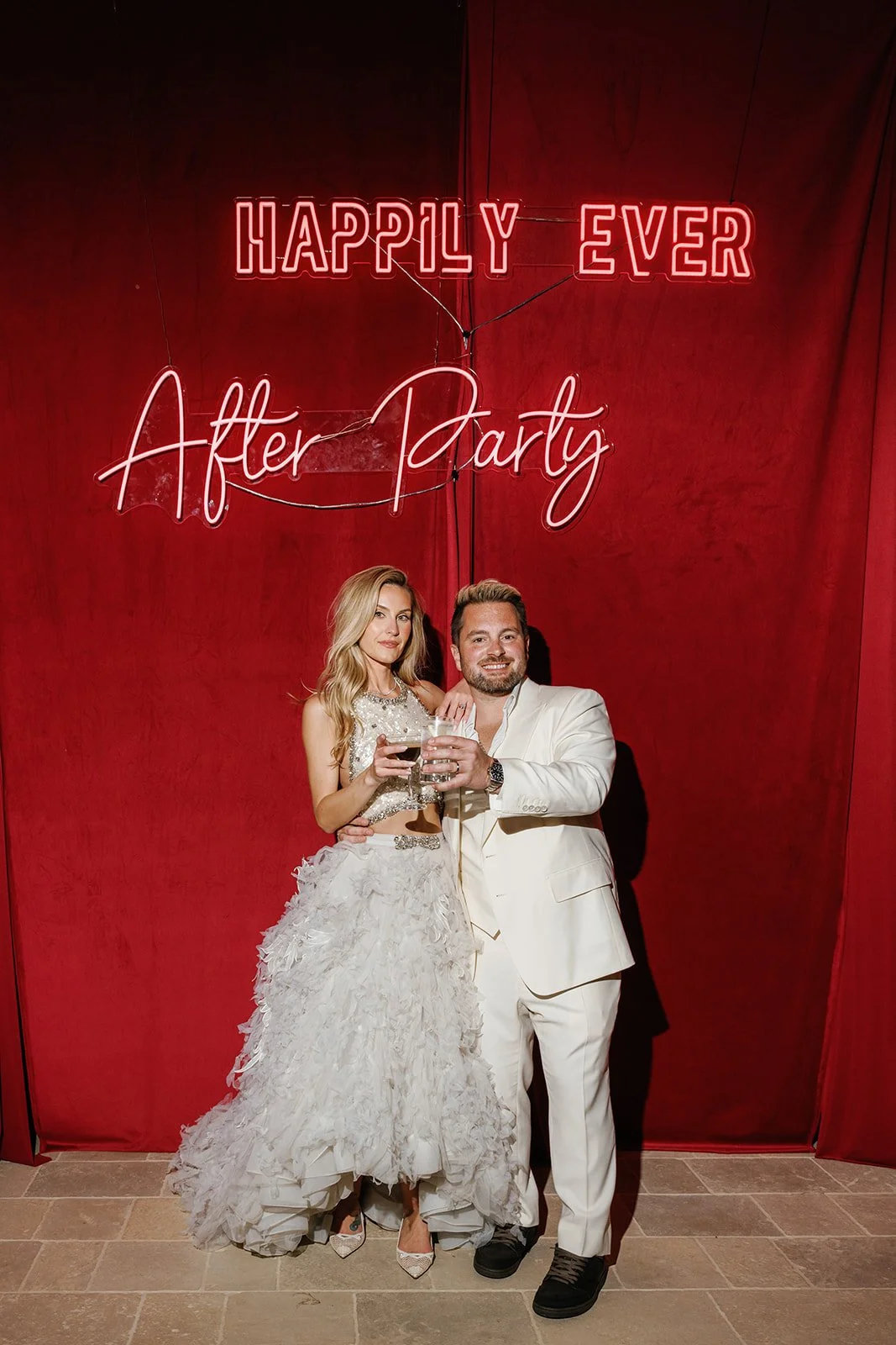 A man and woman in formal attire holding glasses of wine, standing in front of a red backdrop with neon signs that say 'Happily Ever After Party'.