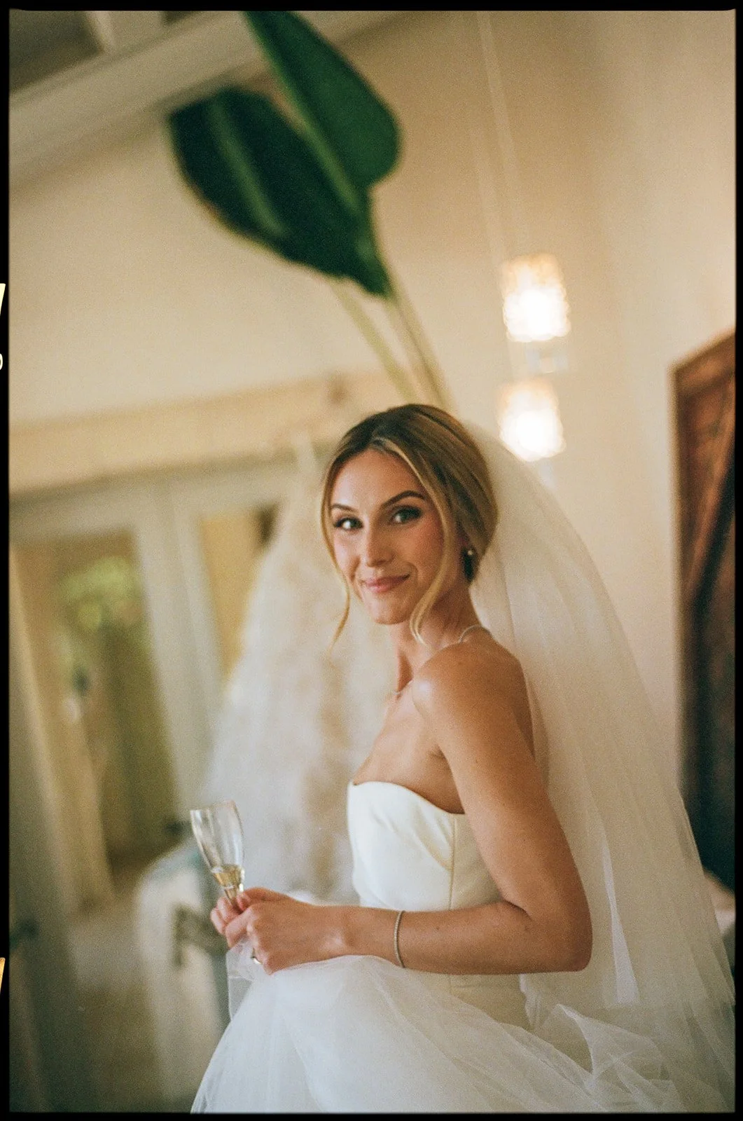 A bride in a strapless white wedding gown holding a champagne flute, smiling at the camera in an indoor setting.