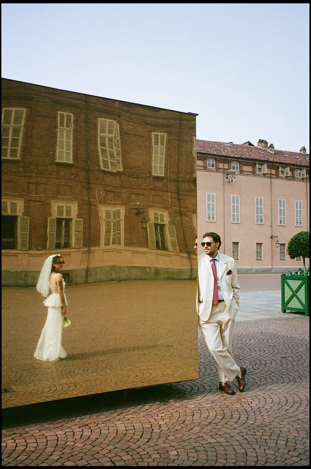 A man in a cream suit and sunglasses leaning against a reflective sculpture, which shows a woman in a white wedding dress and veil, standing on a cobblestone square with historic buildings in the background.