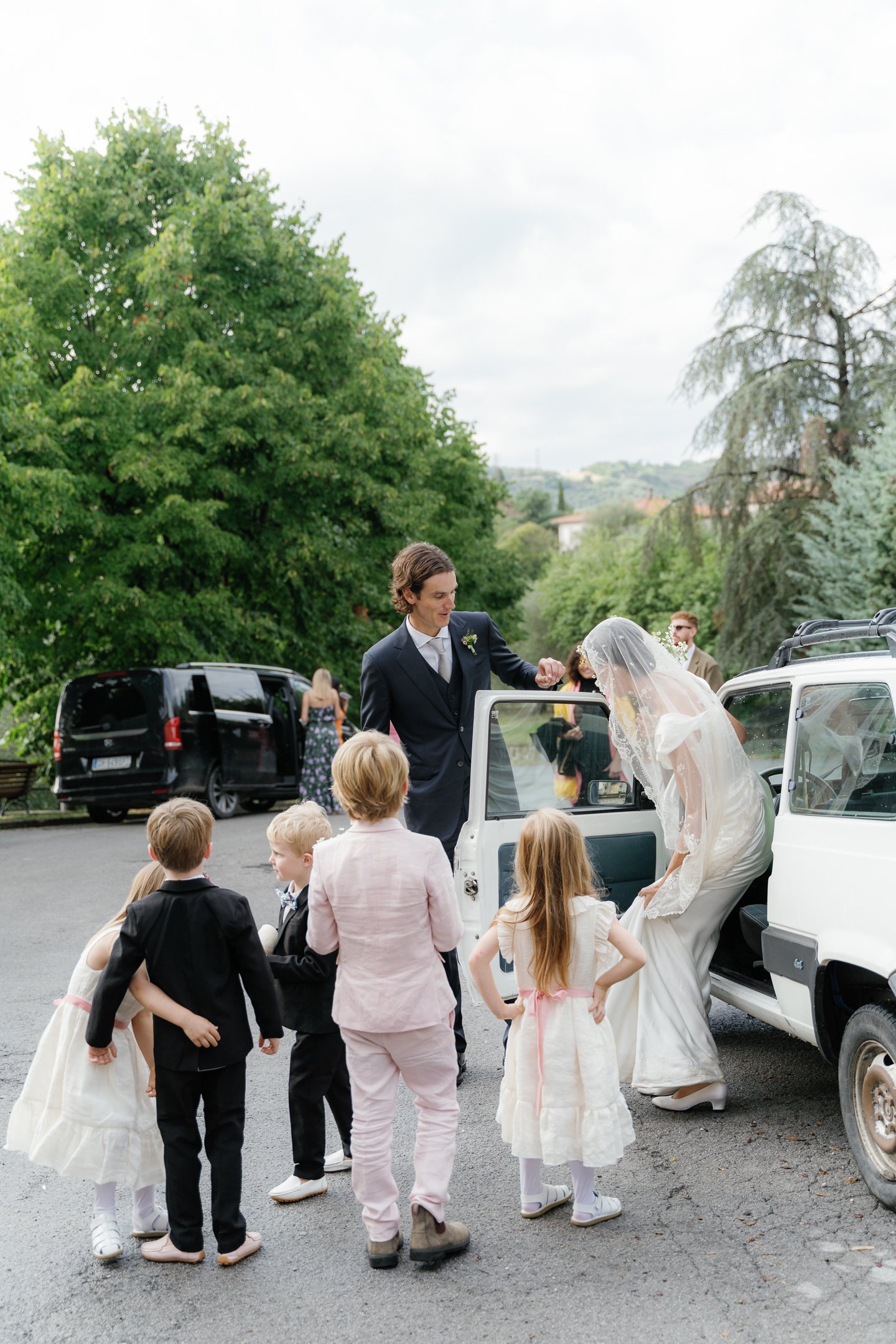 A bride in a white dress and veil getting out of a white car while a groom in a black suit assists her, surrounded by children in formal attire during a wedding event outdoors on a cloudy day.