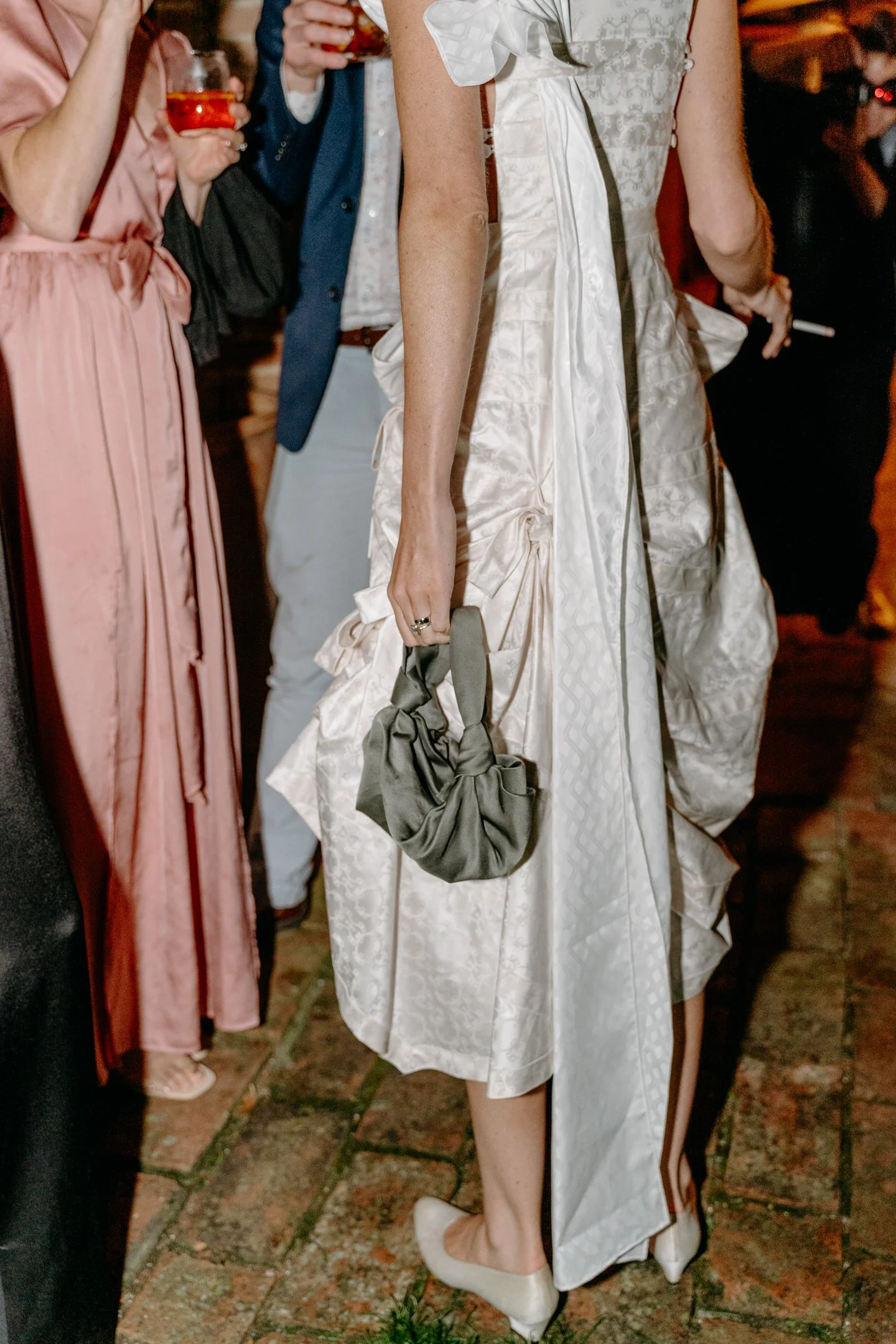 A woman in a white, patterned dress holding a gray, ruched handbag at a social event with other people around, some holding drinks.