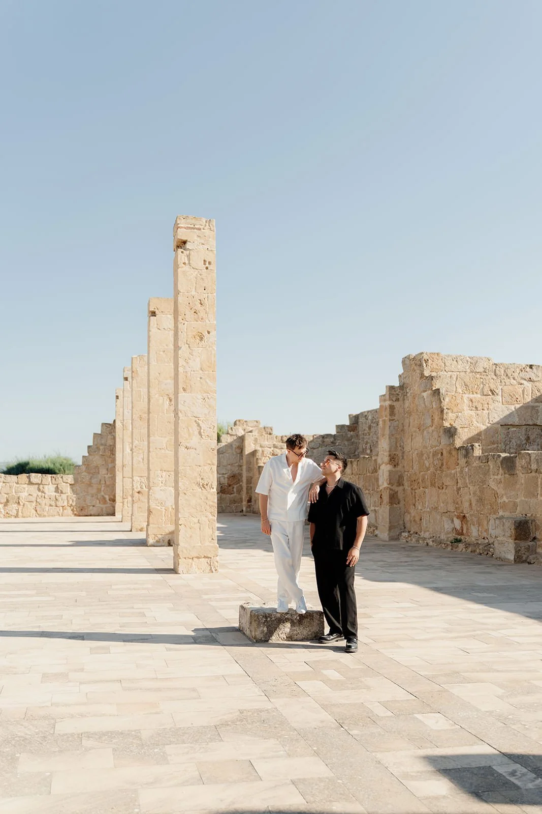 Two people dressed in casual clothing, one in white and one in black, standing among ancient stone ruins with tall columns, under a clear blue sky.
