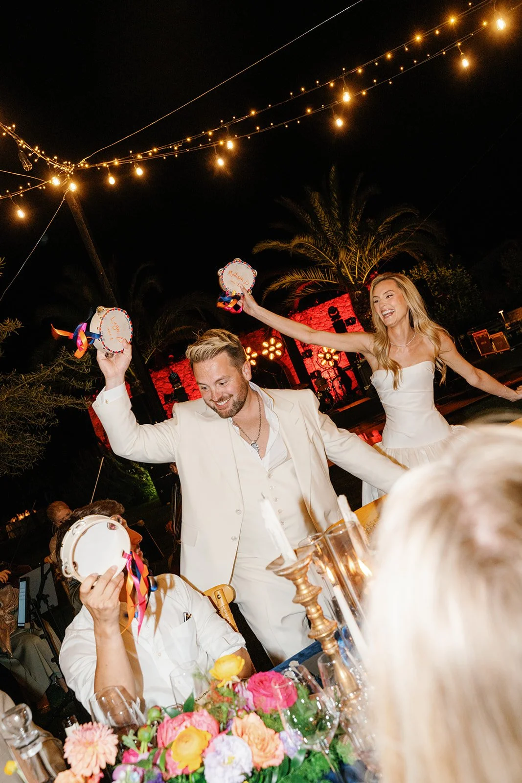 Celebrating at a nighttime outdoor wedding reception with string lights, palm trees, and a stage in the background. The bride and groom are dancing and holding decorated small cakes with ribbons, surrounded by guests at a decorated table with colorfu