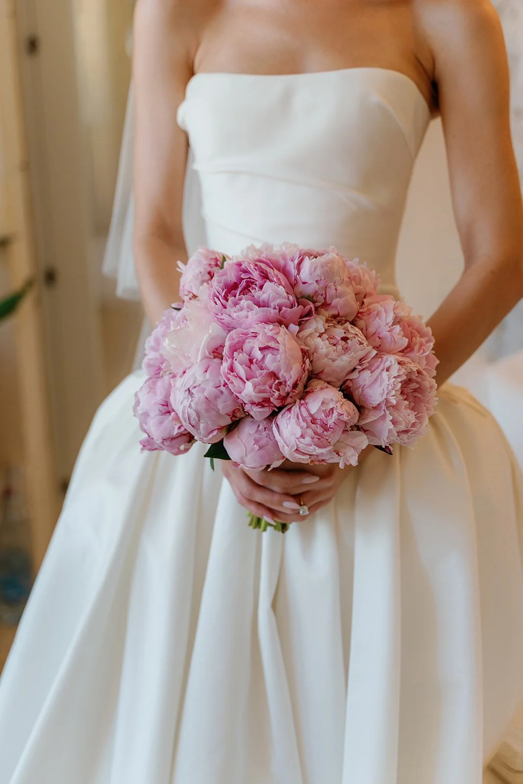 Close-up of a bride holding a bouquet of pink peonies, wearing a strapless white wedding gown.
