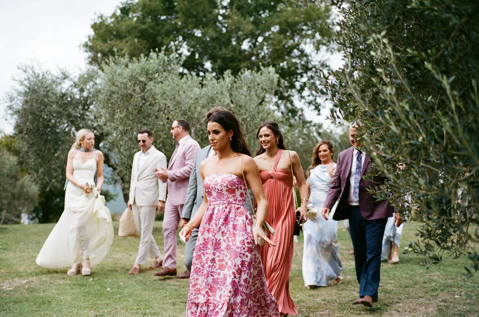 Group of people dressed in elegant formal attire walking outdoors on a grassy path surrounded by trees.
