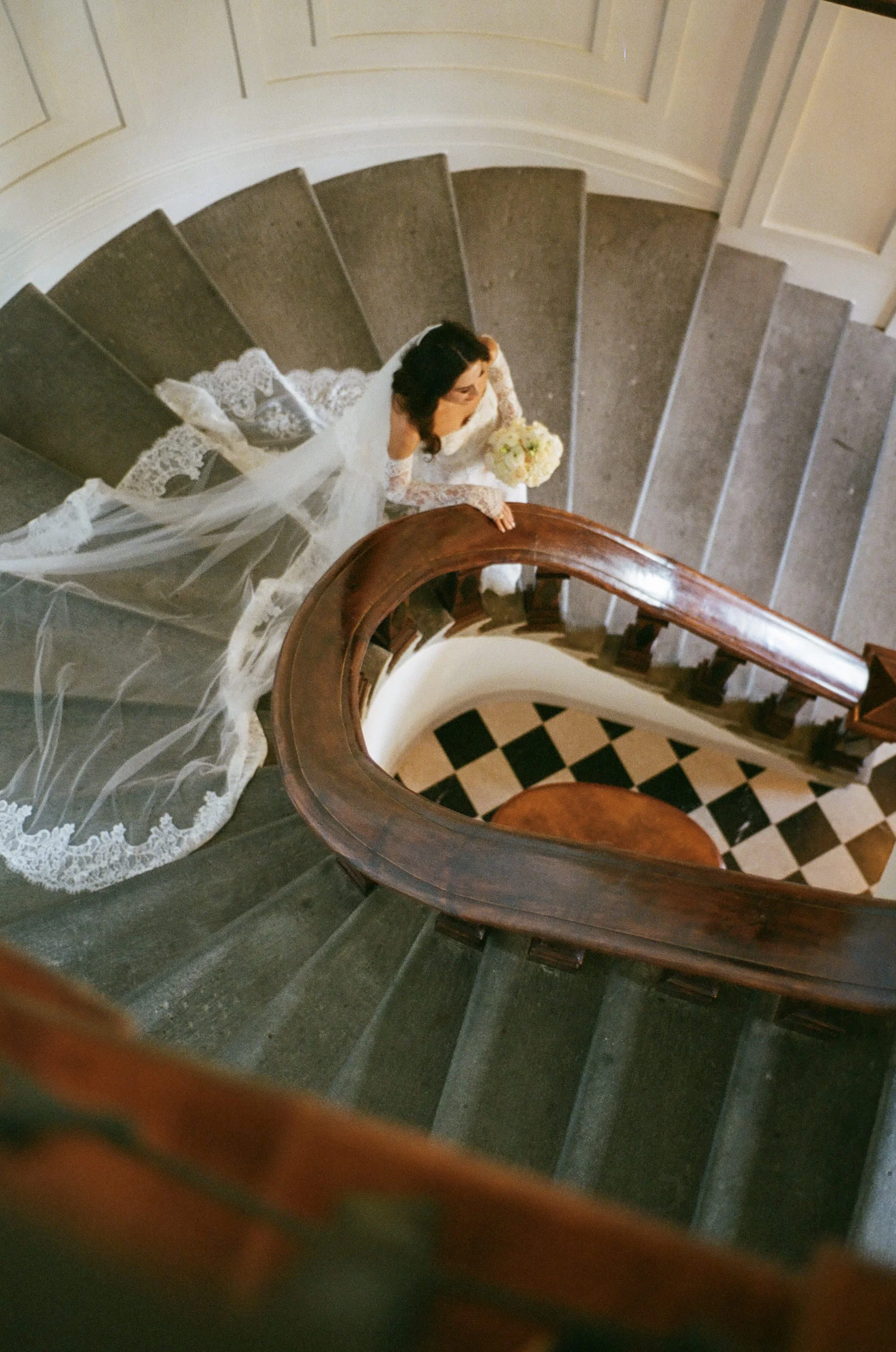 A bride in a white wedding dress with a train is standing at the top of a curved staircase, holding a bouquet, looking up from an overhead perspective.