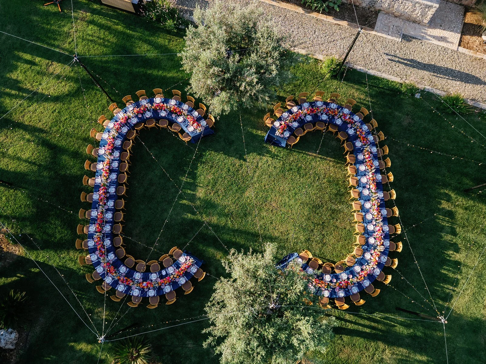 Aerial view of a large outdoor event setting with two long, U-shaped tables decorated with colorful floral centerpieces, set on a grassy lawn with a few picnic tables and trees nearby.