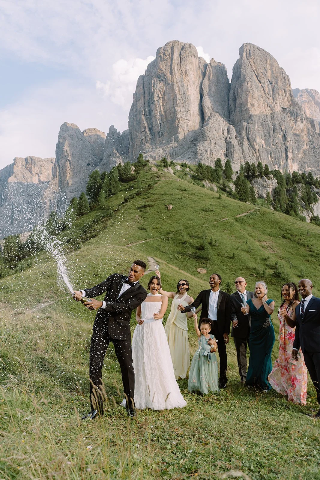 A wedding celebration outdoors with a group of people, including a bride and groom, standing on a grassy hillside with mountains in the background. The groom is spraying champagne, and everyone is smiling and enjoying the moment.