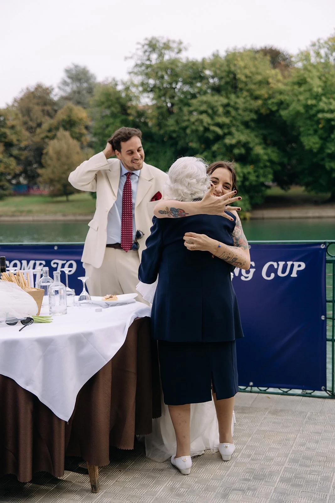 Two women hug and smile as a man looks on happily, behind a table with wine glasses and a water bottle near a lake with trees.