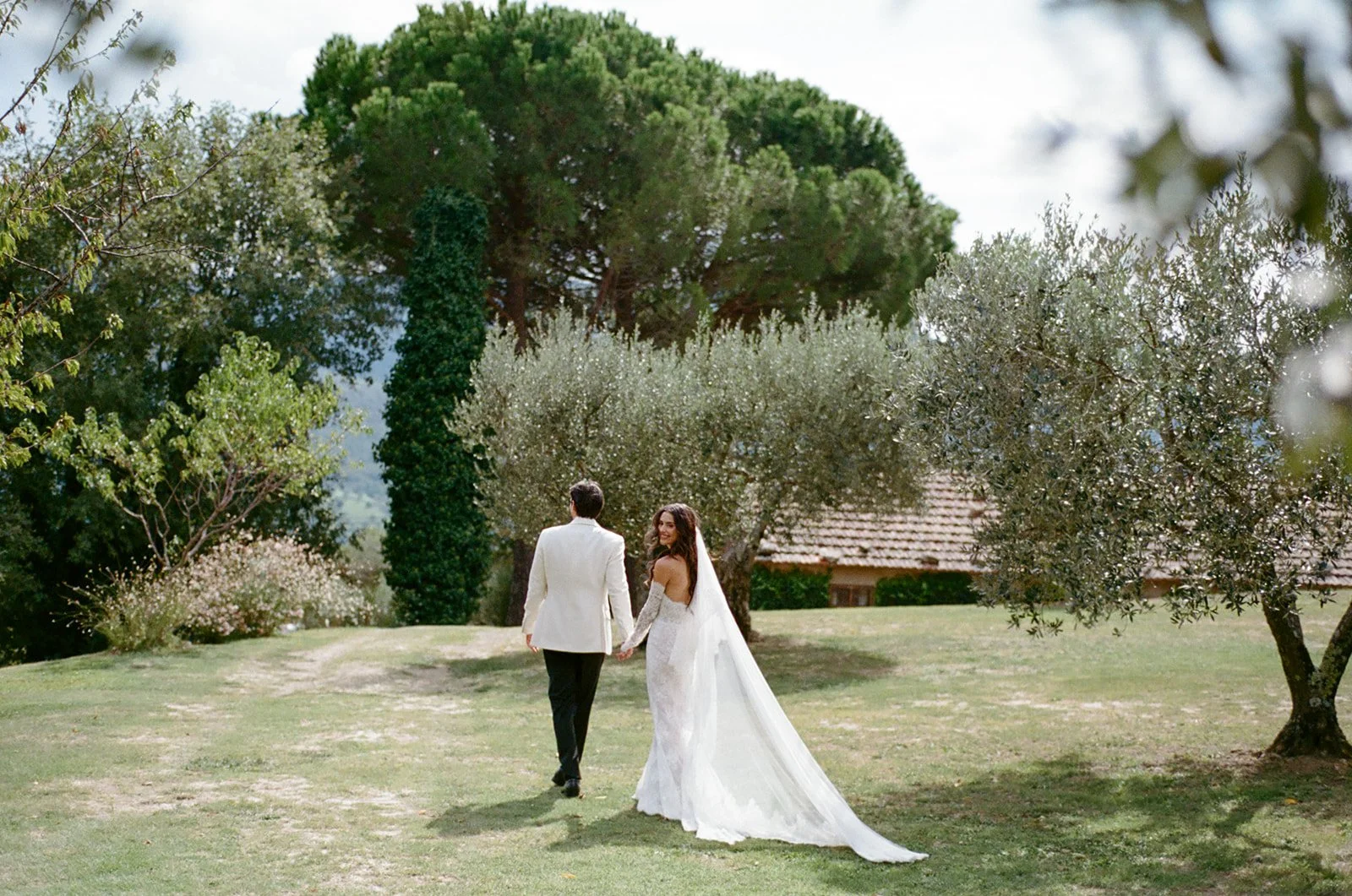 A bride and groom holding hands, walking away in a garden setting on their wedding day, surrounded by trees and greenery.