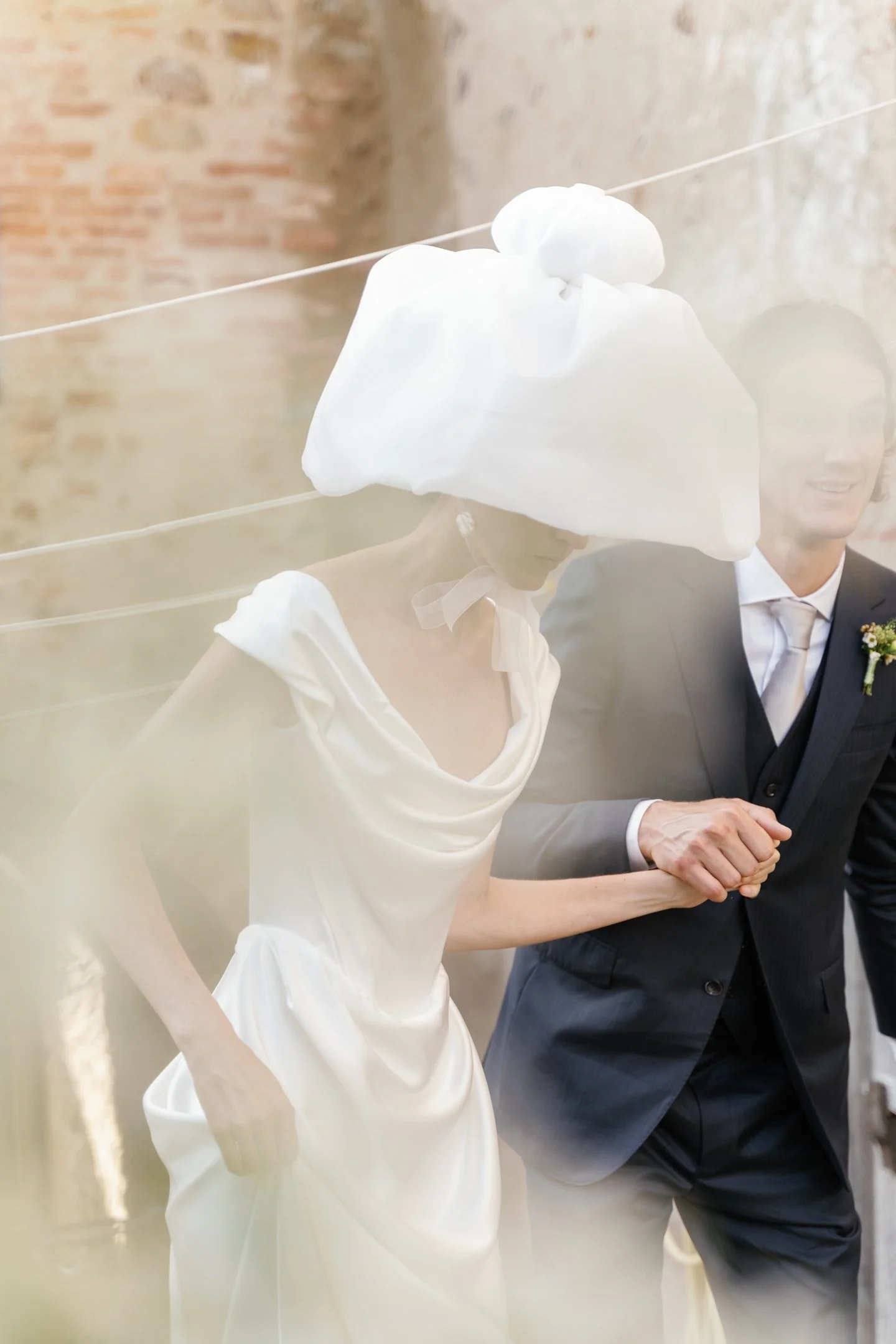 A bride and groom holding hands during their wedding ceremony, with the bride wearing a white satin dress and a large white hat, and the groom in a dark suit with a boutonniere, in front of a rustic brick wall.