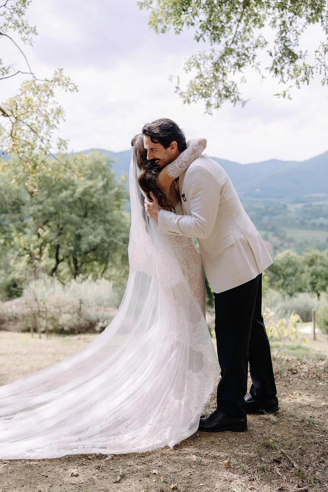 A bride and groom are hugging outdoors against a scenic background of trees and mountains, on a cloudy day. The bride wears a white lace wedding dress with a long train, and the groom wears a white suit jacket and black trousers.