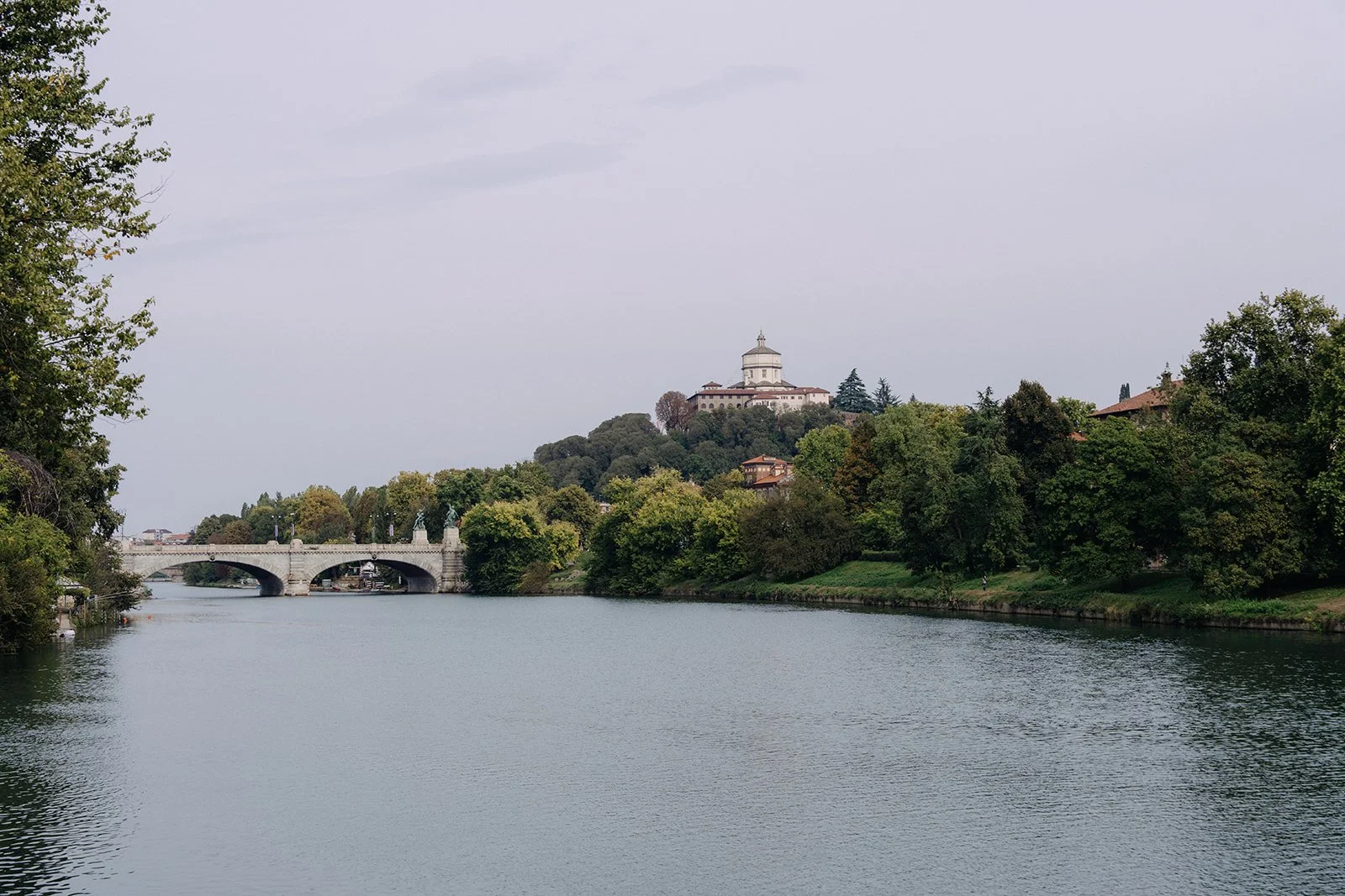 View of a river with a bridge on the left and a hill with a large white building at the top on the right, surrounded by green trees.