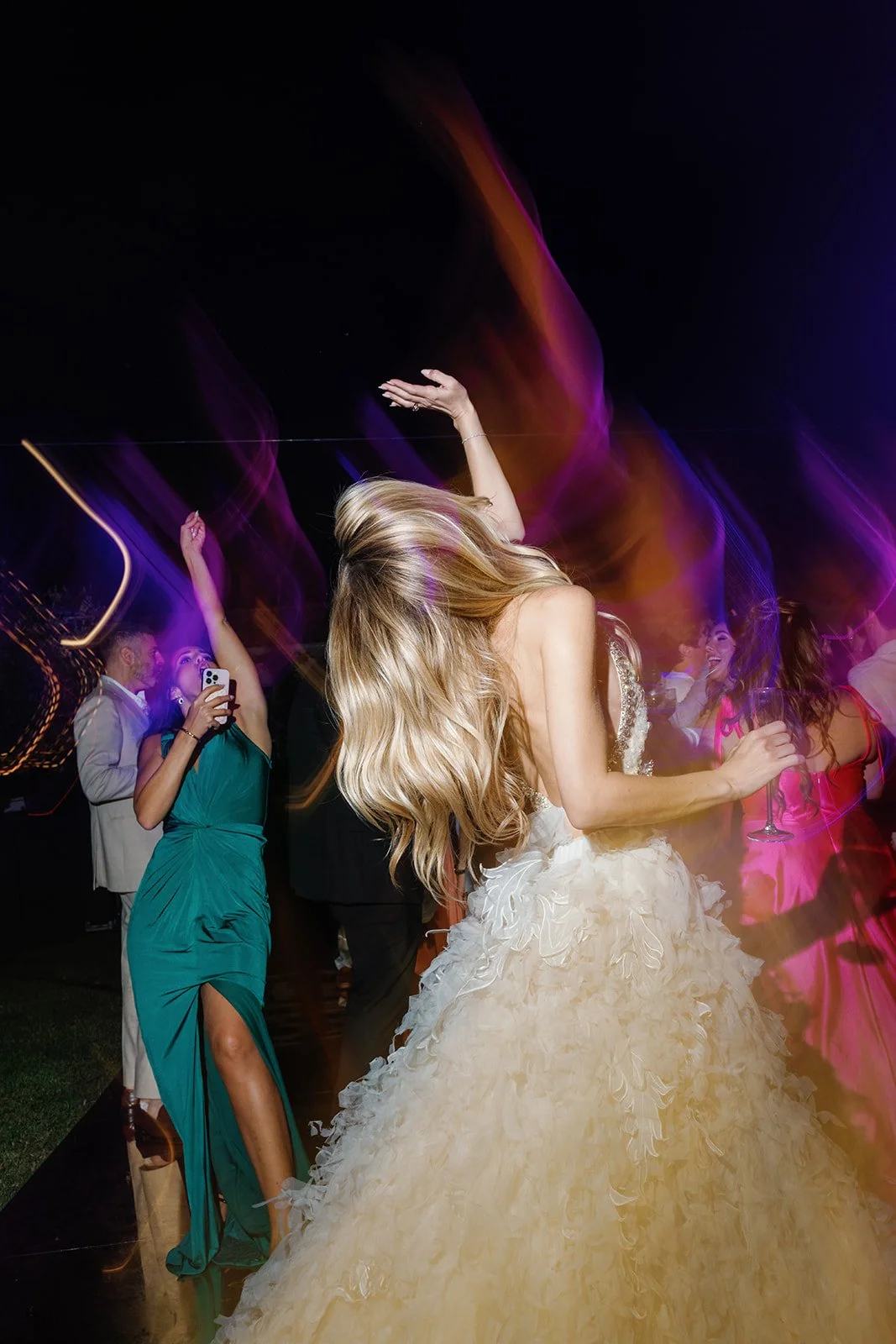 A woman in a white wedding dress dancing at a party with others, colorful light trails around her, at night.
