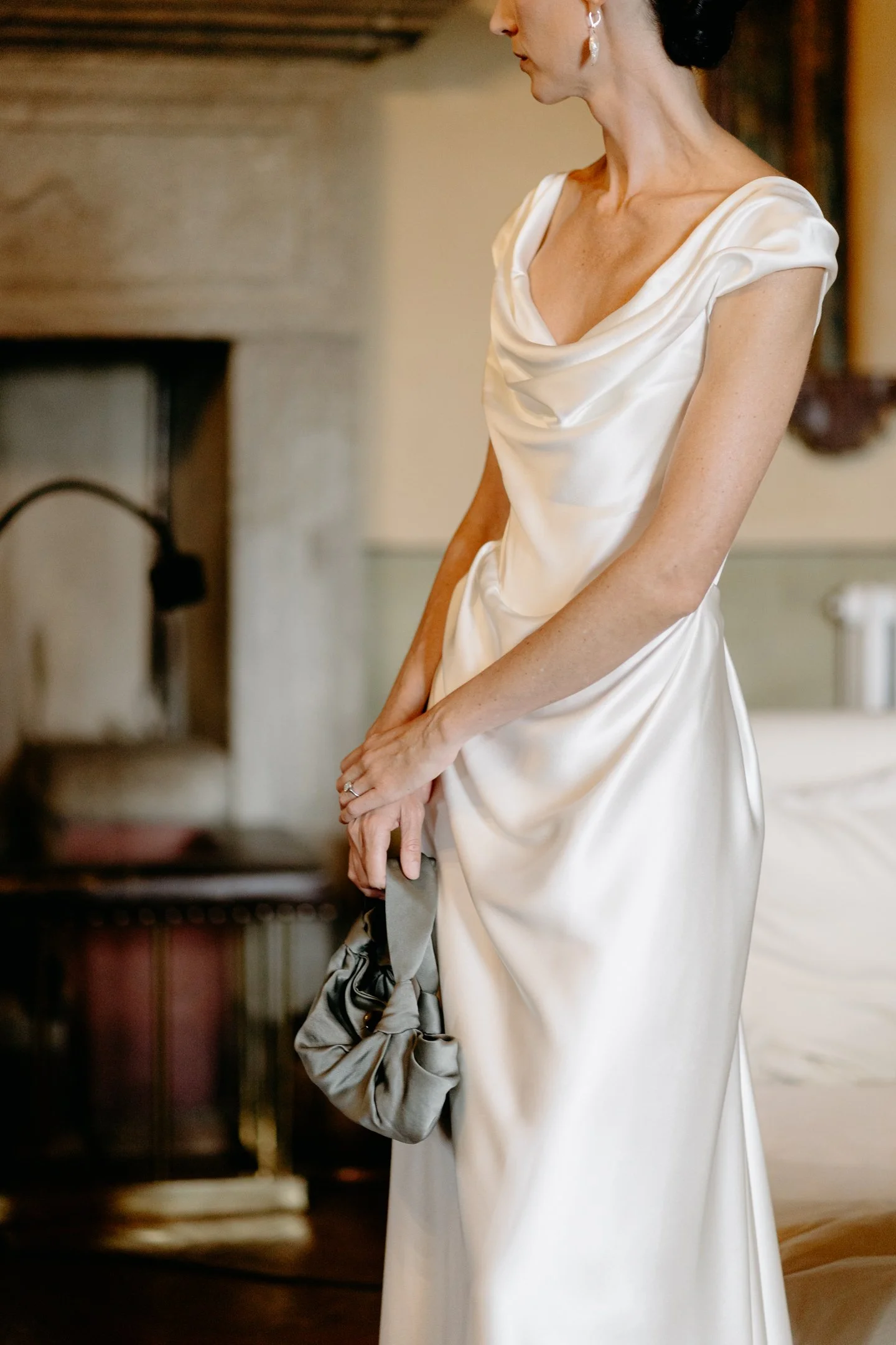 A woman in an elegant white satin dress holding a small gray clutch bag, standing indoors near a bed, with a focus on her upper body and side profile.