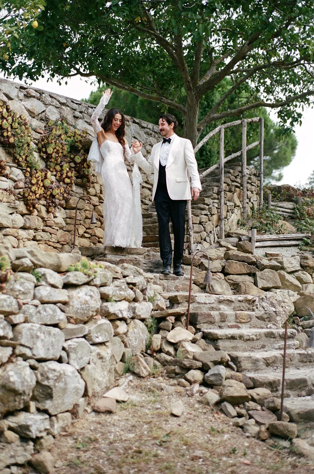 A bride and groom walking down stone steps outdoors, holding hands, celebrating, with a tree and stone wall in the background.