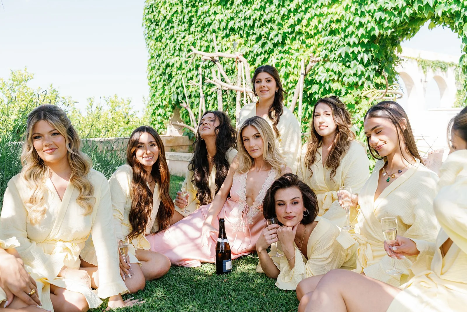 A group of women having a picnic outdoors, sitting on grass with drinks, in front of a lush, green ivy-covered wall.