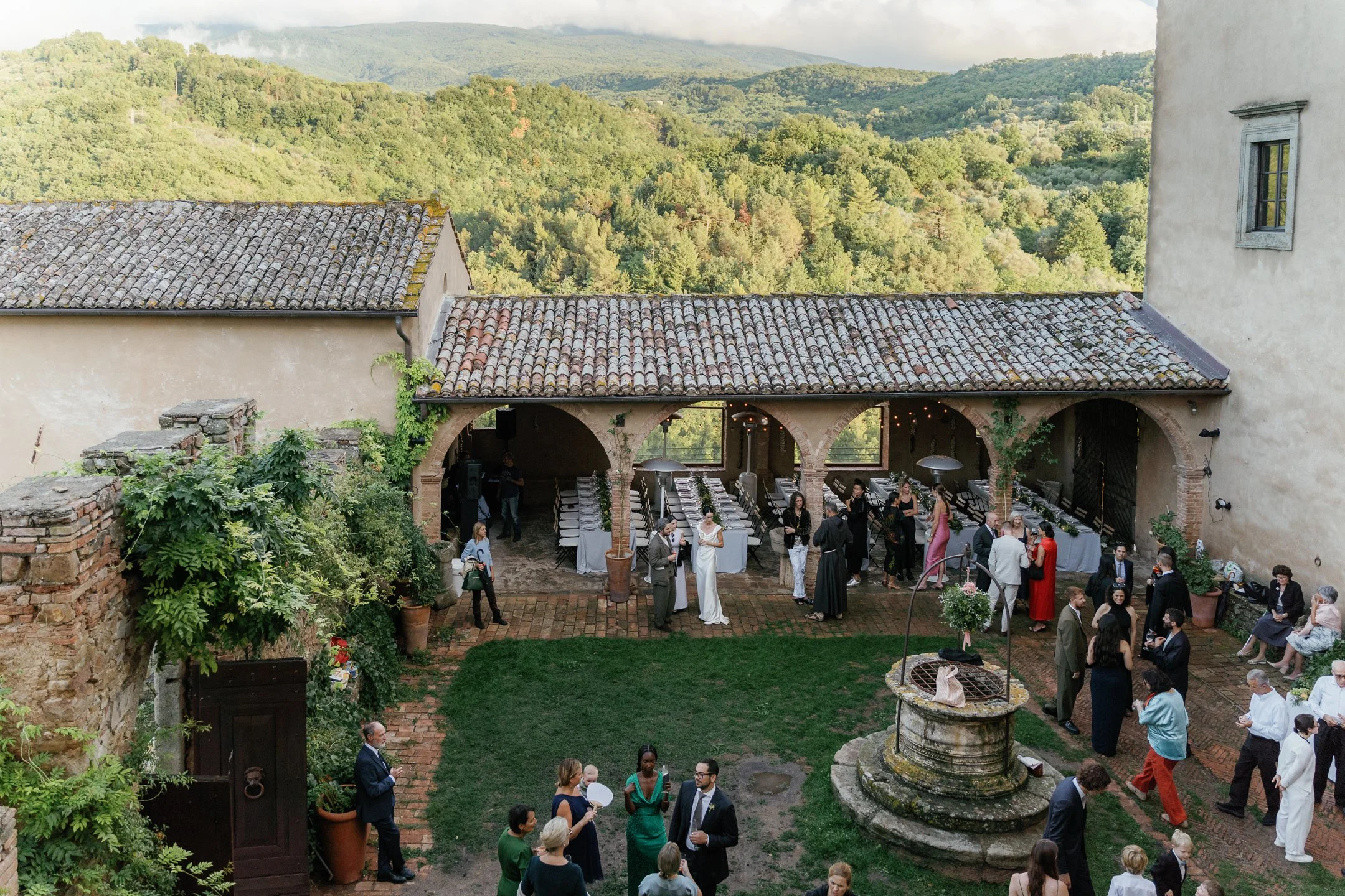 People gathered at an outdoor wedding reception in a courtyard with a well, surrounded by lush green hills and rustic buildings.